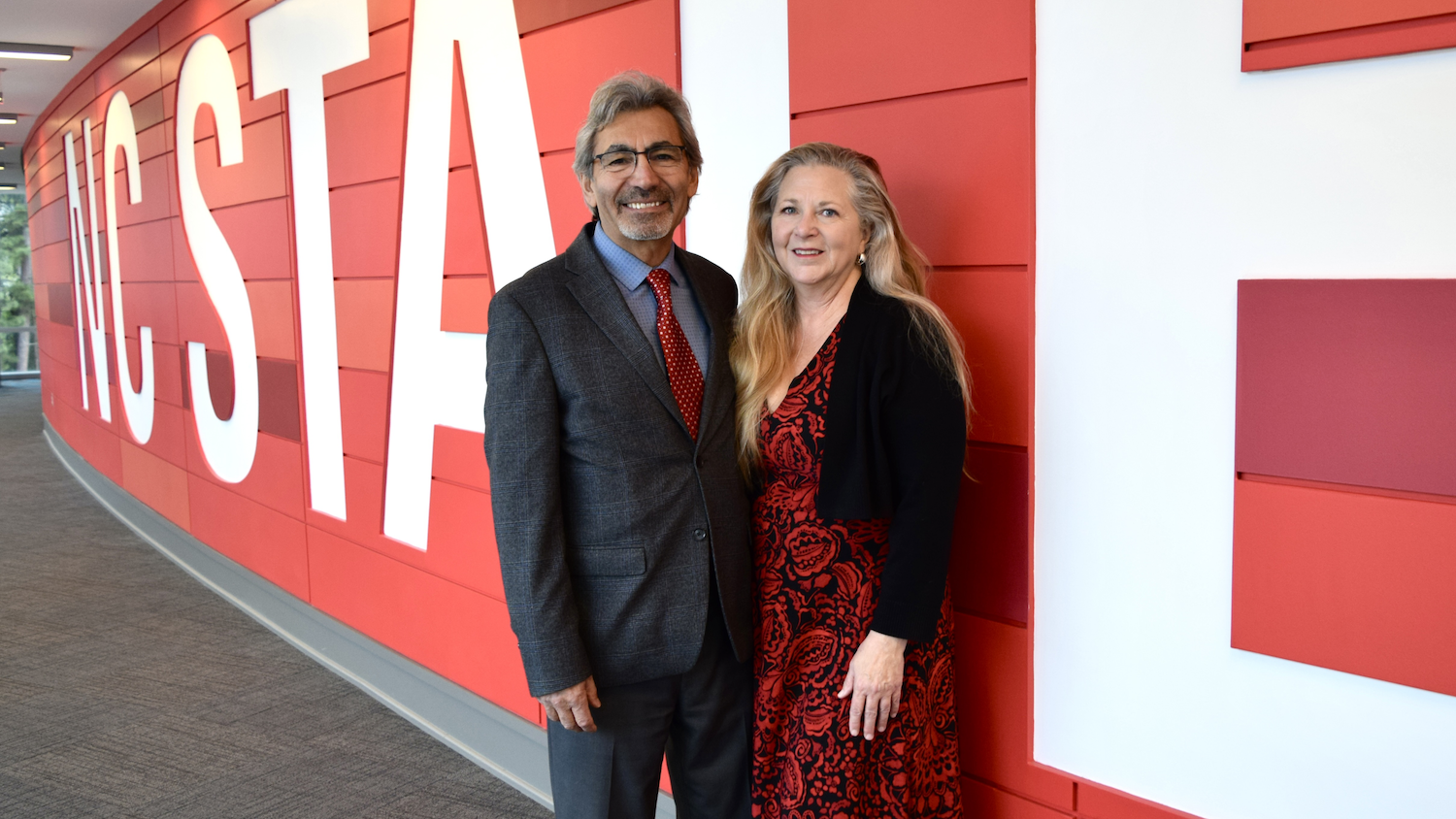 A man and woman standing together in front of a red wall with large white letters spelling 'HISTORICAL'. The man is wearing glasses, a gray suit, light blue shirt, and red tie. The woman has long blonde hair and is wearing a red and black patterned dress with a black blazer.