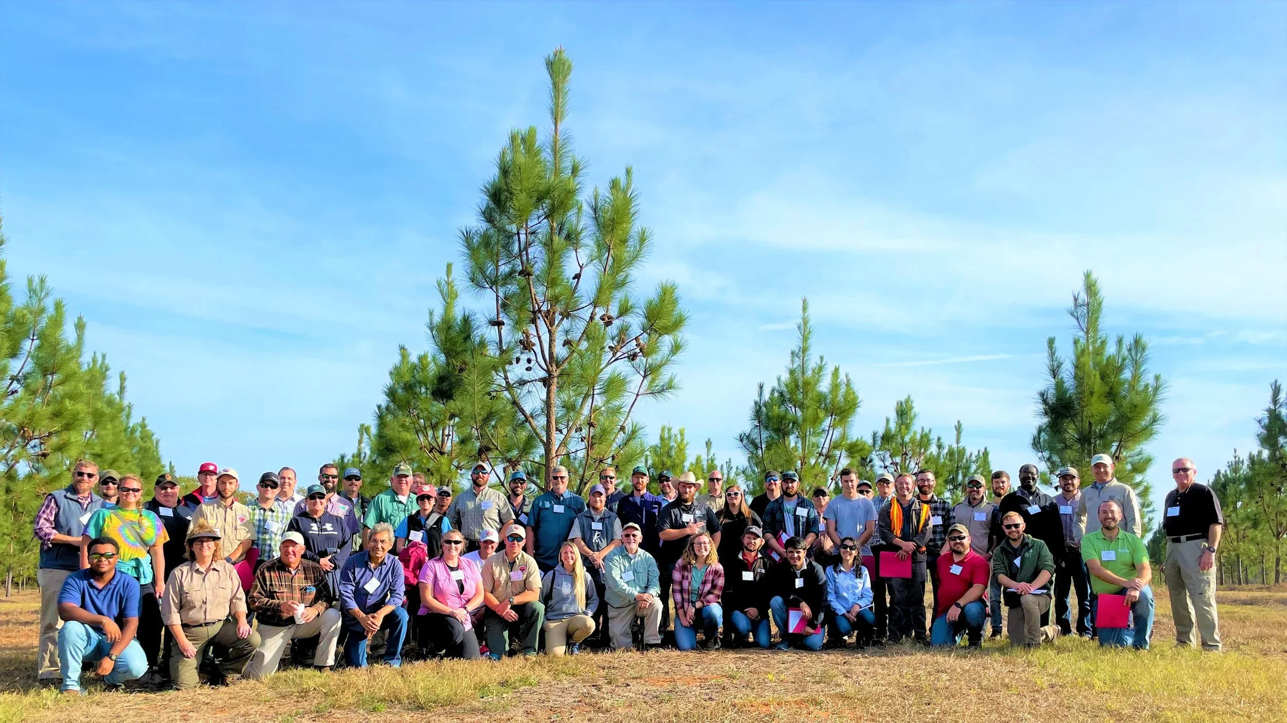 A large group of people outdoors in front of pine trees under a blue sky, posing for a group photo.