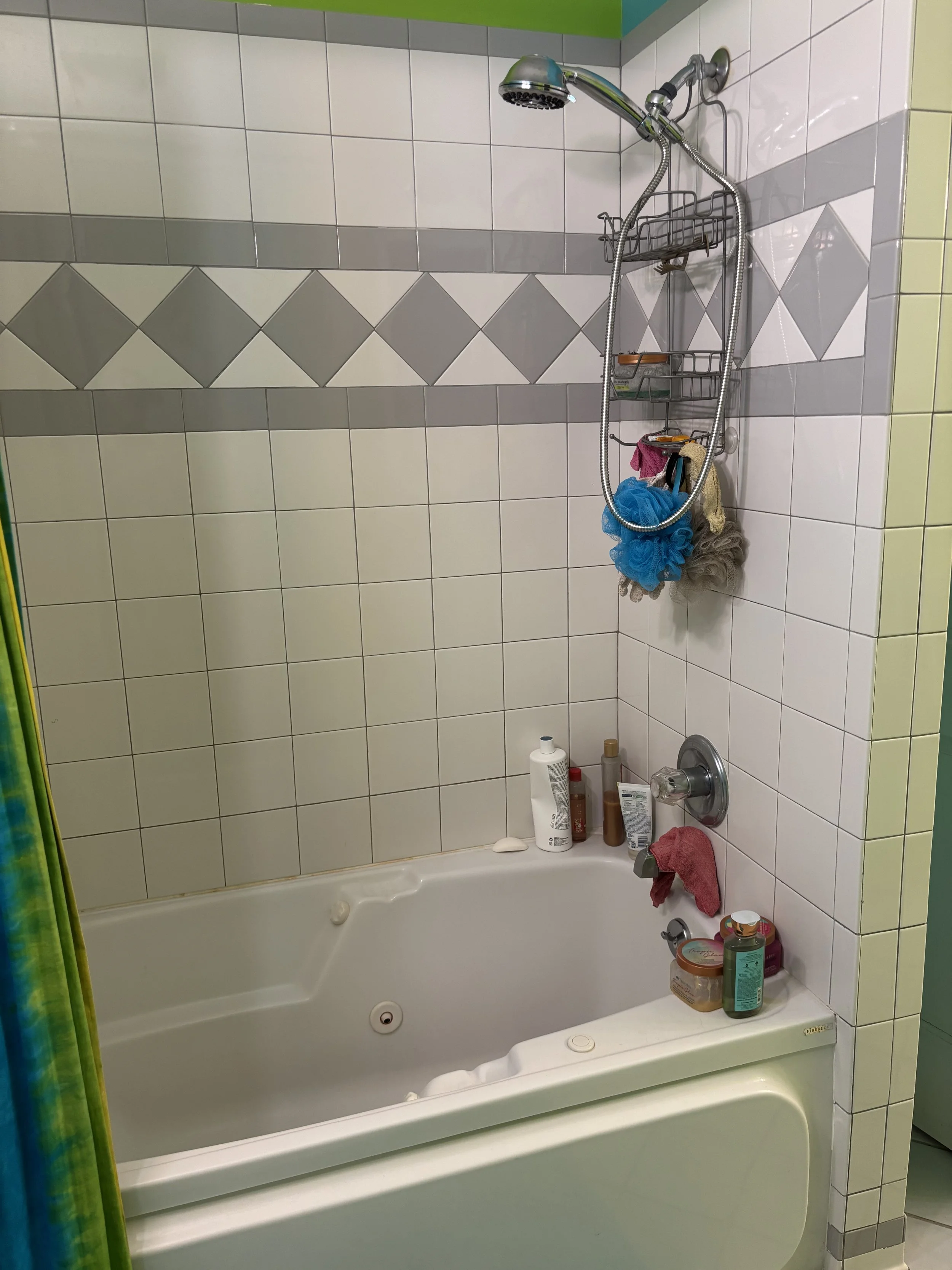 A bathroom with a bathtub, tiled walls, and shower fixtures. Several bottles of toiletries, a towel, and cleaning supplies are on the edge of the tub. A shower caddy with additional items hangs from the showerhead.