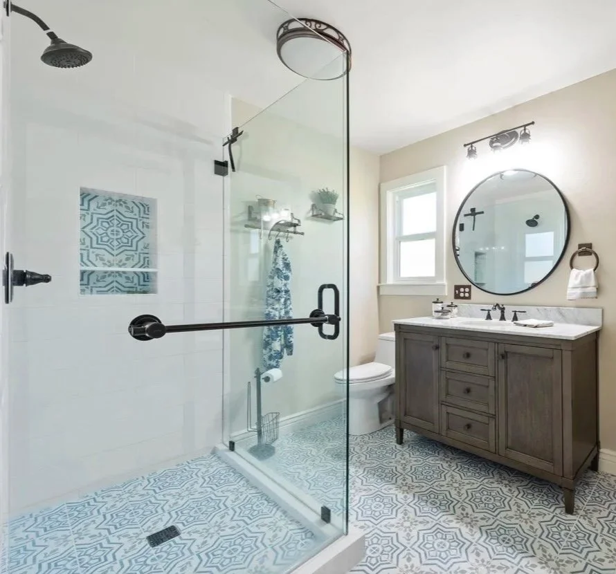 Modern bathroom with walk-in glass shower, round mirror above wooden vanity, black fixtures, patterned floor tiles, and a small window.