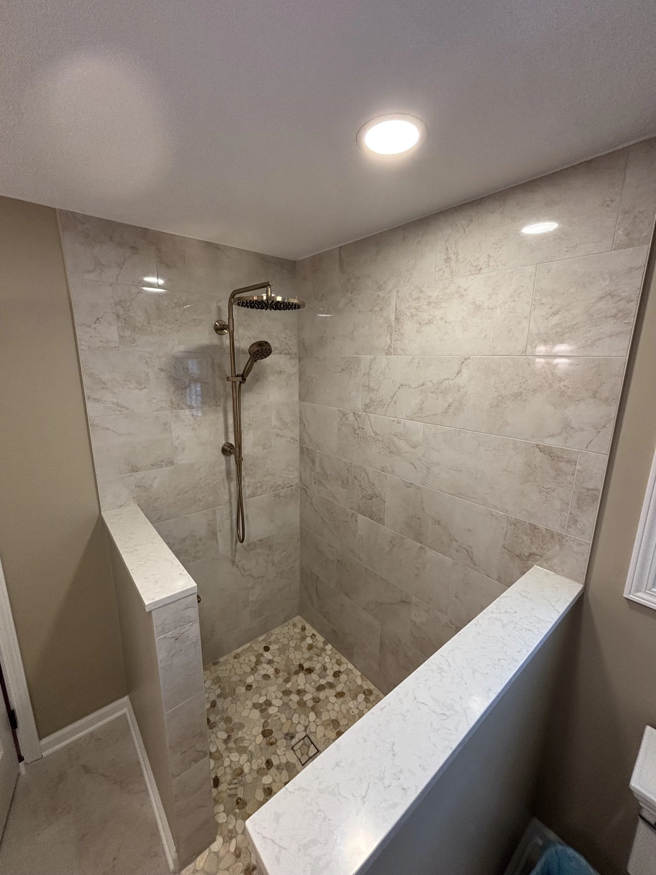 A walk-in shower with beige marble tiles, a pebble stone floor, a stainless steel showerhead, and a partial wall divider.