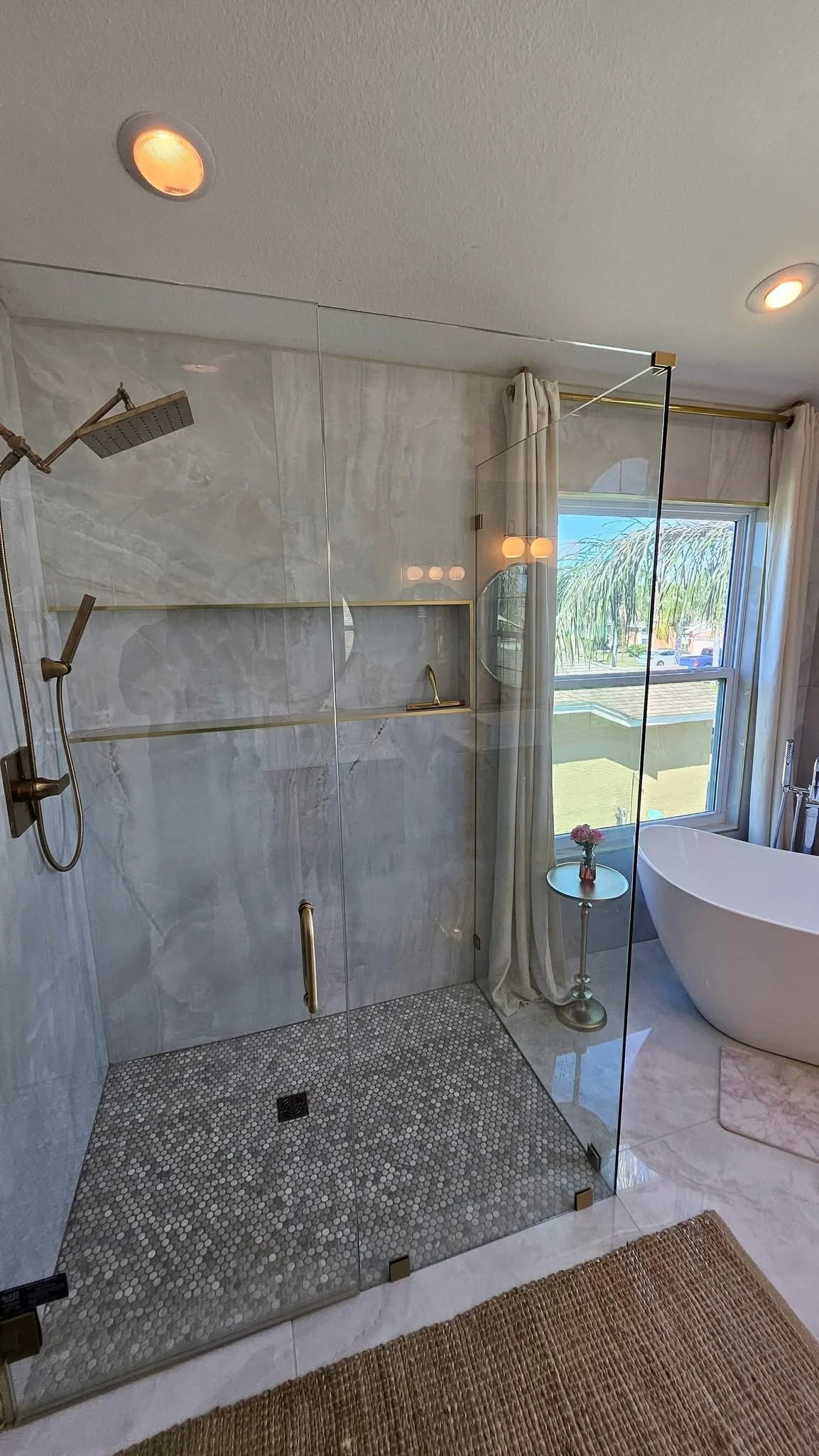 A modern bathroom with a walk-in shower enclosed with glass, featuring two shower heads, a white freestanding bathtub near a large window with curtains, a small round table with flowers, and a textured brown rug.