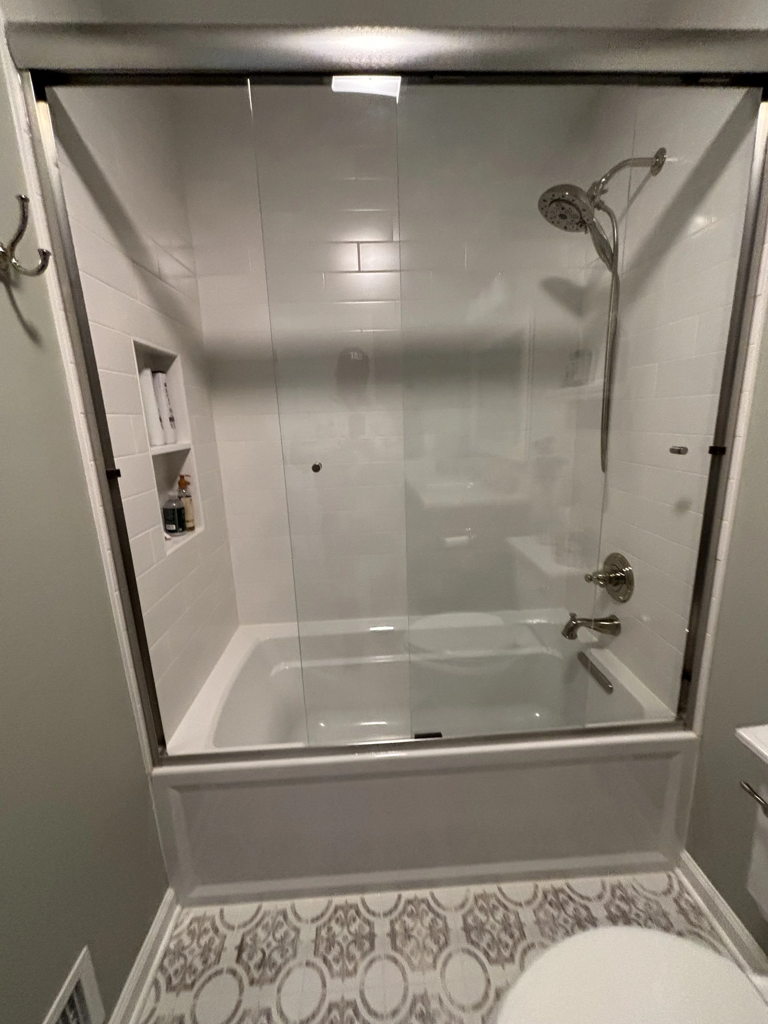 Bathroom shower area with glass door, white subway tile walls, a built-in shelf with toiletries, a rainfall showerhead, and patterned floor tiles.