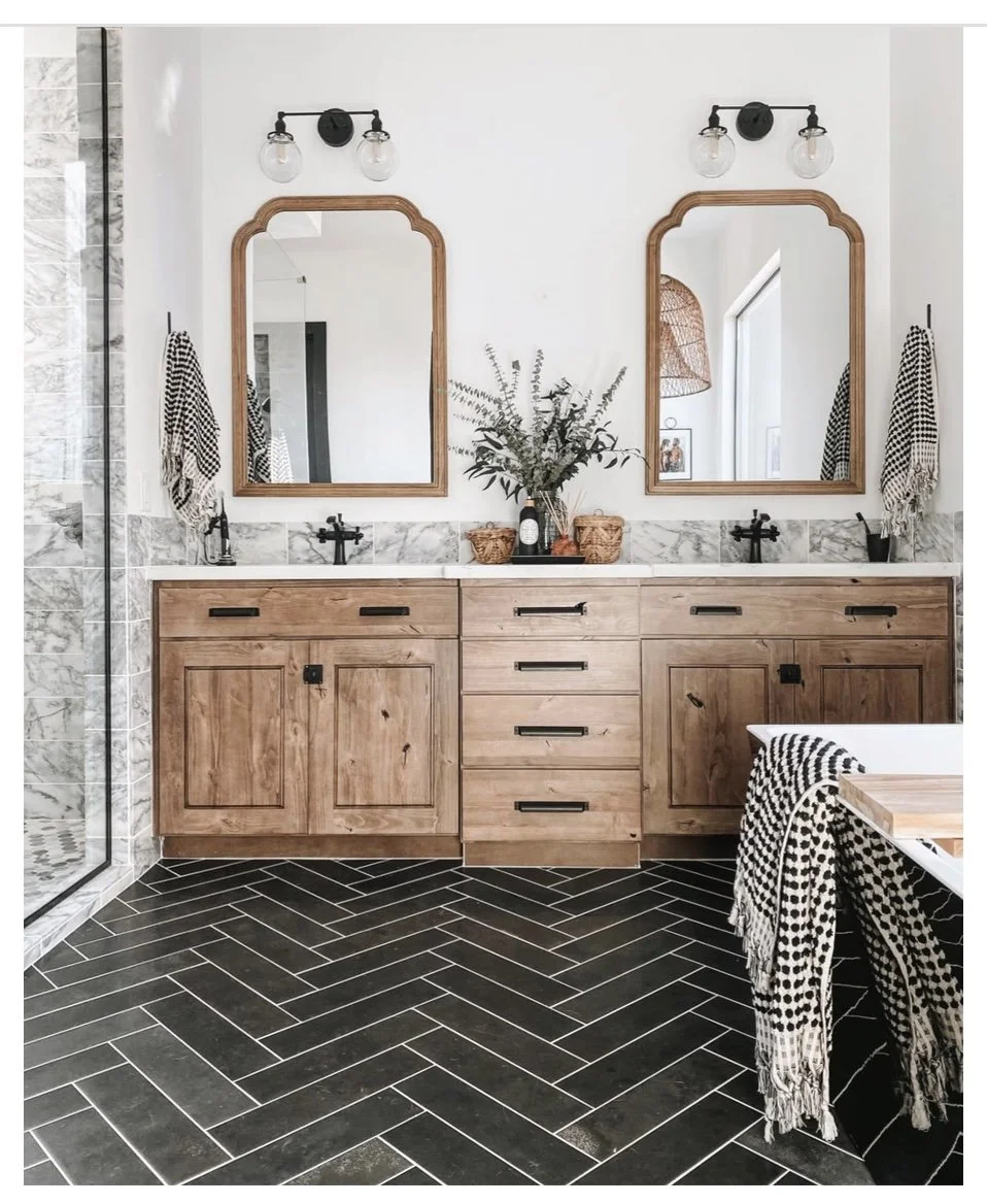 A modern bathroom with a double vanity featuring natural wood cabinets, black hardware, and two wooden framed mirrors. The wall above the vanity has two light fixtures with exposed bulbs, and there are black-and-white patterned towels hanging on each side. The countertop has a large vase with greenery and two small wicker baskets. The floor is covered with black herringbone tiles, and part of a glass shower with stone tiles is visible on the left.
