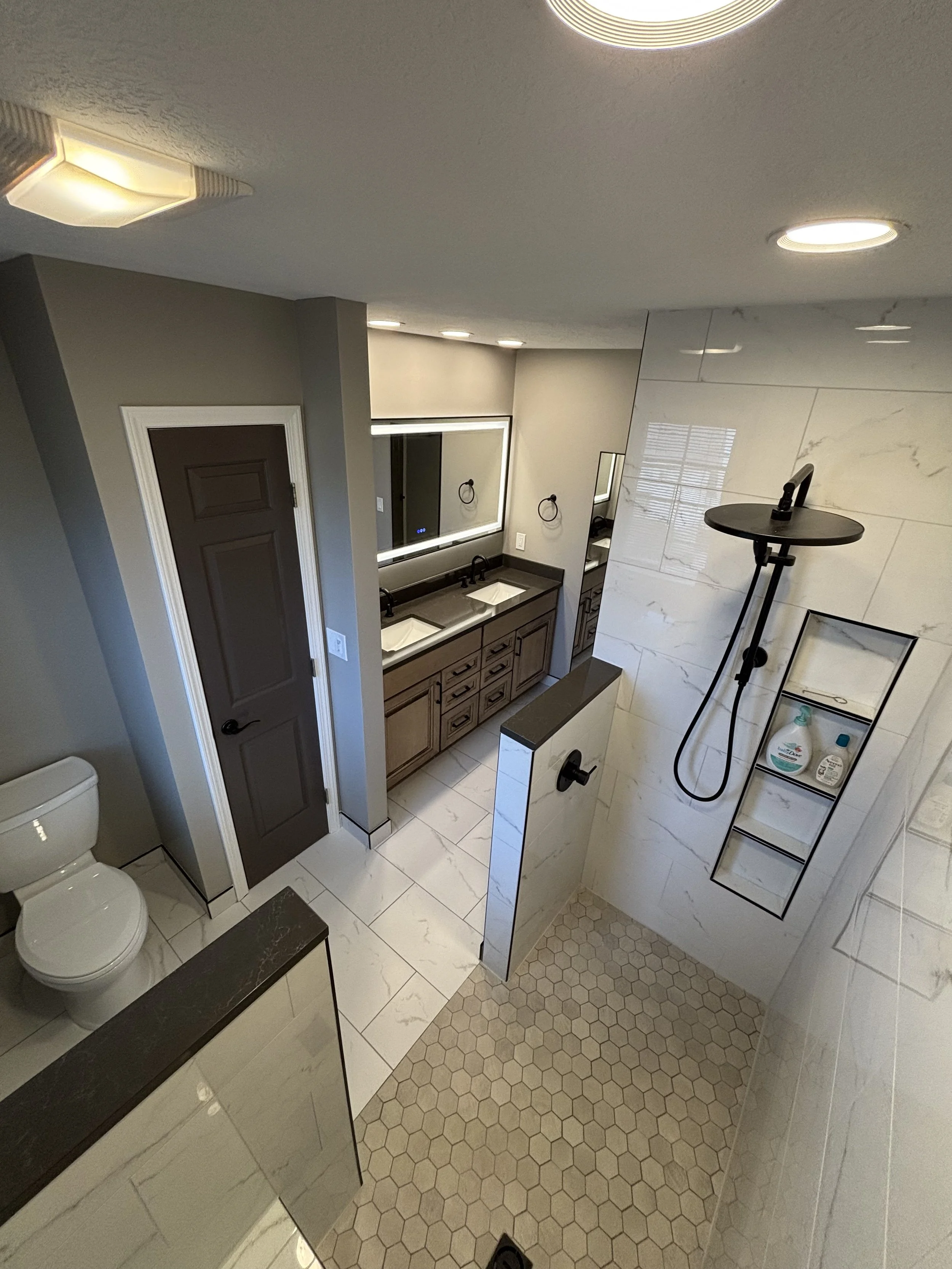 A modern bathroom with a walk-in shower, double sink vanity, toilet, and a mirror with LED lighting. The shower area has a black showerhead and a built-in shelf with toiletries. The room features tile floors and walls with a mix of white marble and hexagonal tiles.