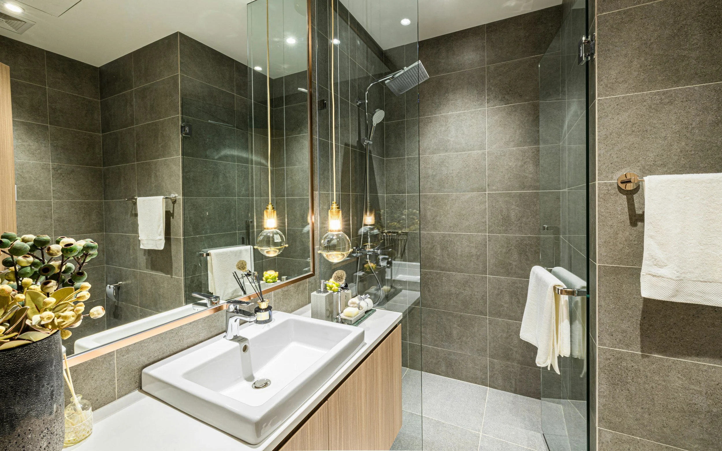 Modern bathroom with dark tiled walls, a white sink, a large mirror, pendant lights, and a glass-enclosed shower area with a rain showerhead, towels on a rack, and decorative plants.