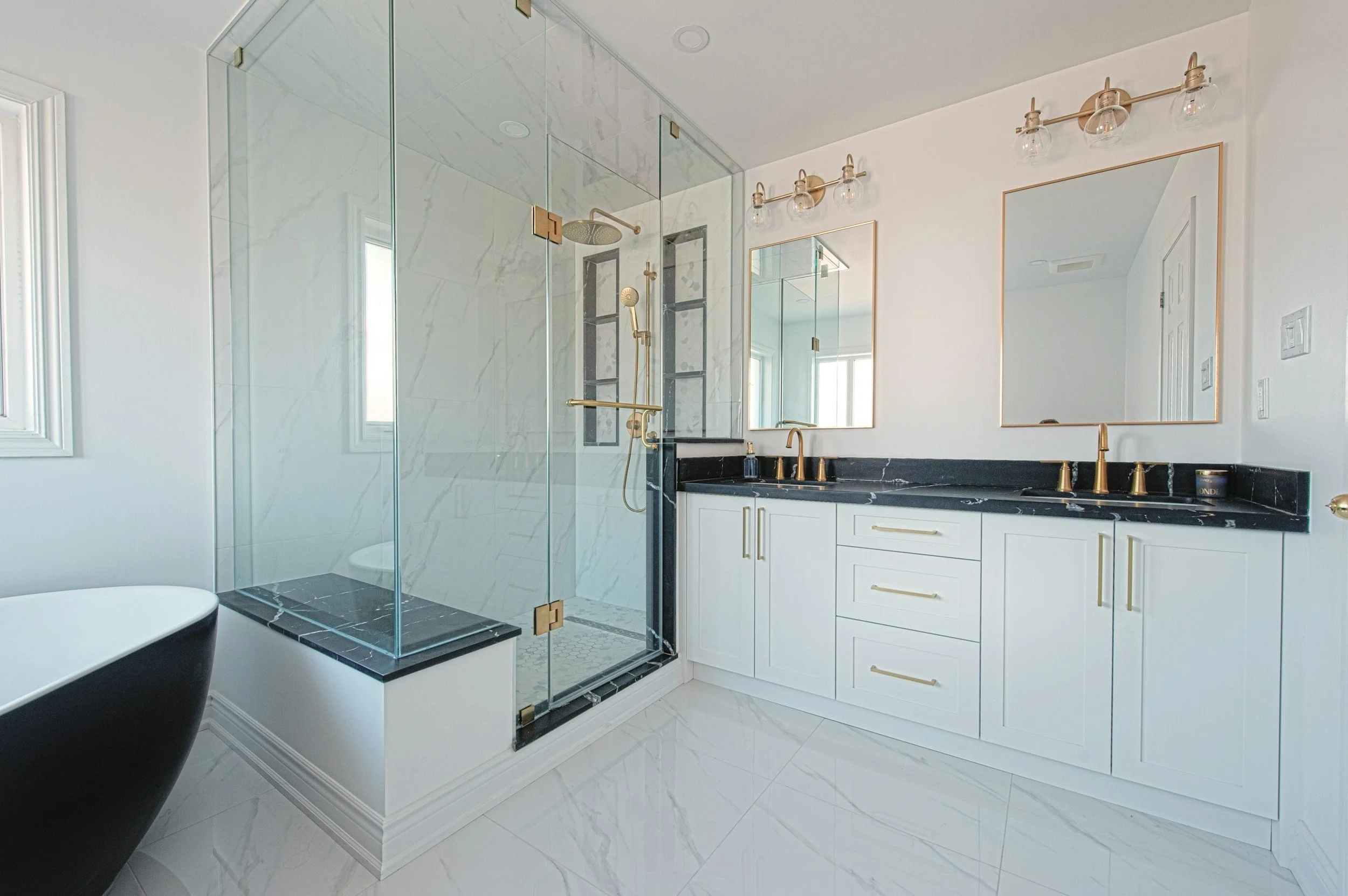 Modern bathroom with glass-enclosed shower, black marble countertops, and white cabinets with gold hardware.