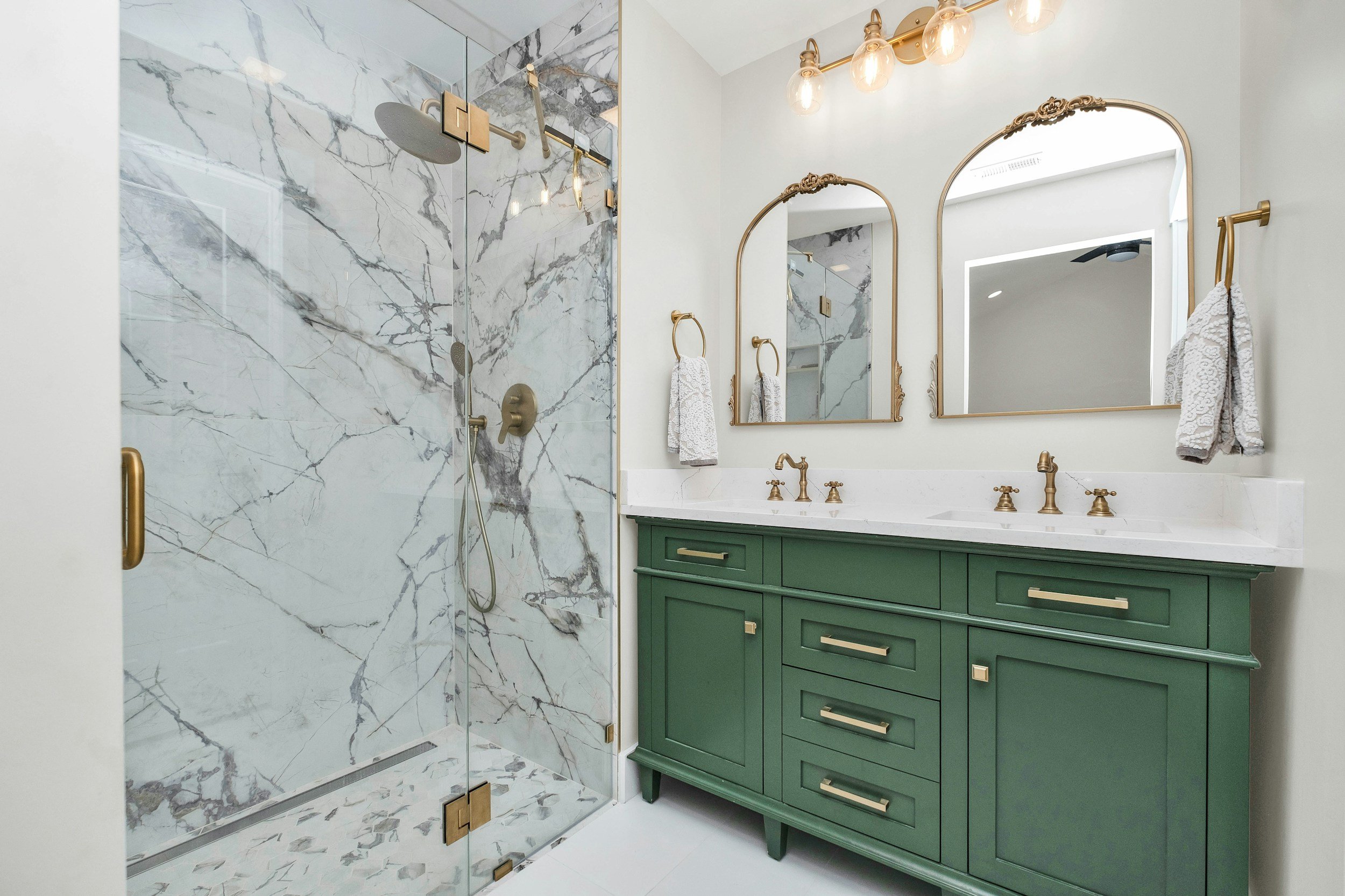 Modern bathroom with a shower featuring marble tiles, green vanity with gold hardware, large mirror with gold frame, and a white countertop with two sinks.