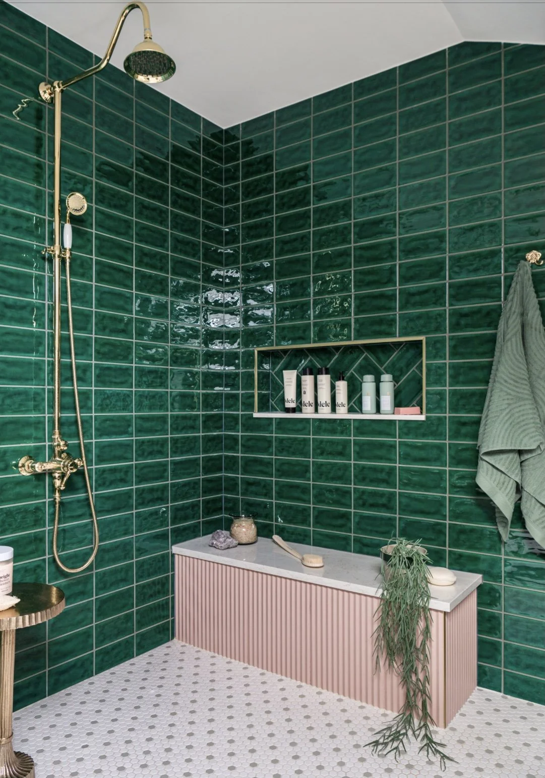 A bathroom with glossy dark green tiled walls, a white ceiling, and a black and white hexagonal patterned floor. It features a gold showerhead and fixtures, a built-in wall shelf with toiletries, a pink-taped bench with a jar, rock, brush, and a plant hanging over the edge, and a green towel hanging on the right wall.