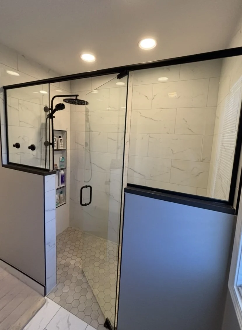 A modern walk-in shower with a black-framed glass enclosure, white marble tiles on the walls, and hexagonal tiles on the floor. It features a black rainfall showerhead and built-in shelves for toiletries.