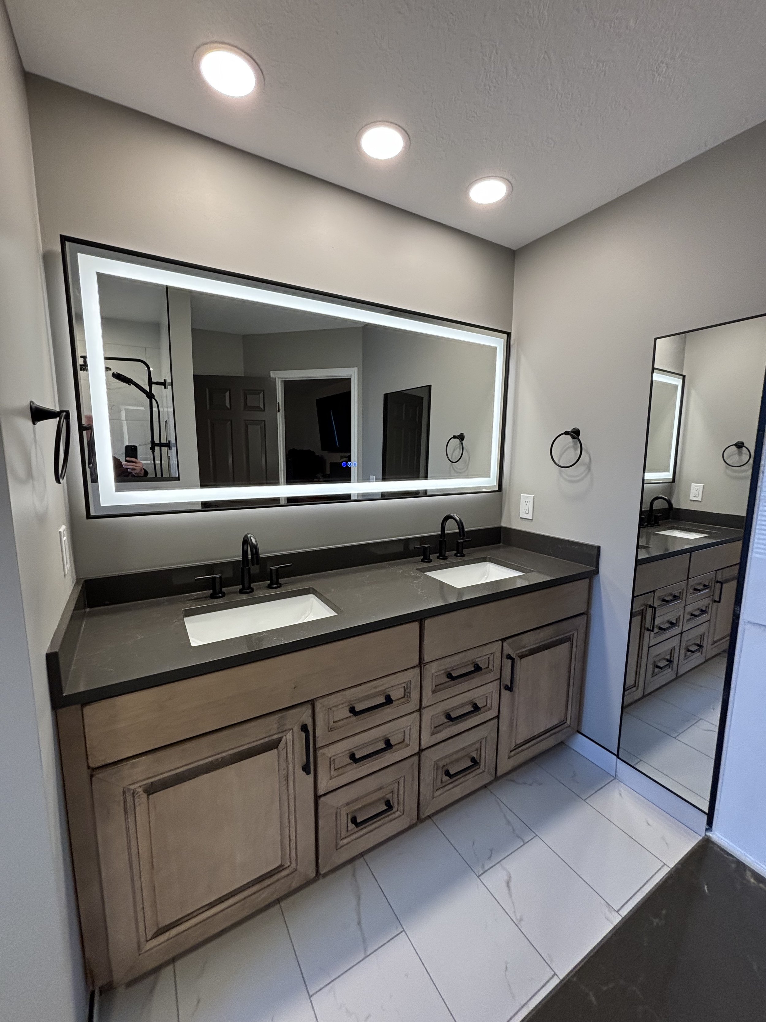 Bathroom vanity with a large illuminated mirror, dual sinks with black faucets, wooden cabinetry, and a mirror door leading to a shower.