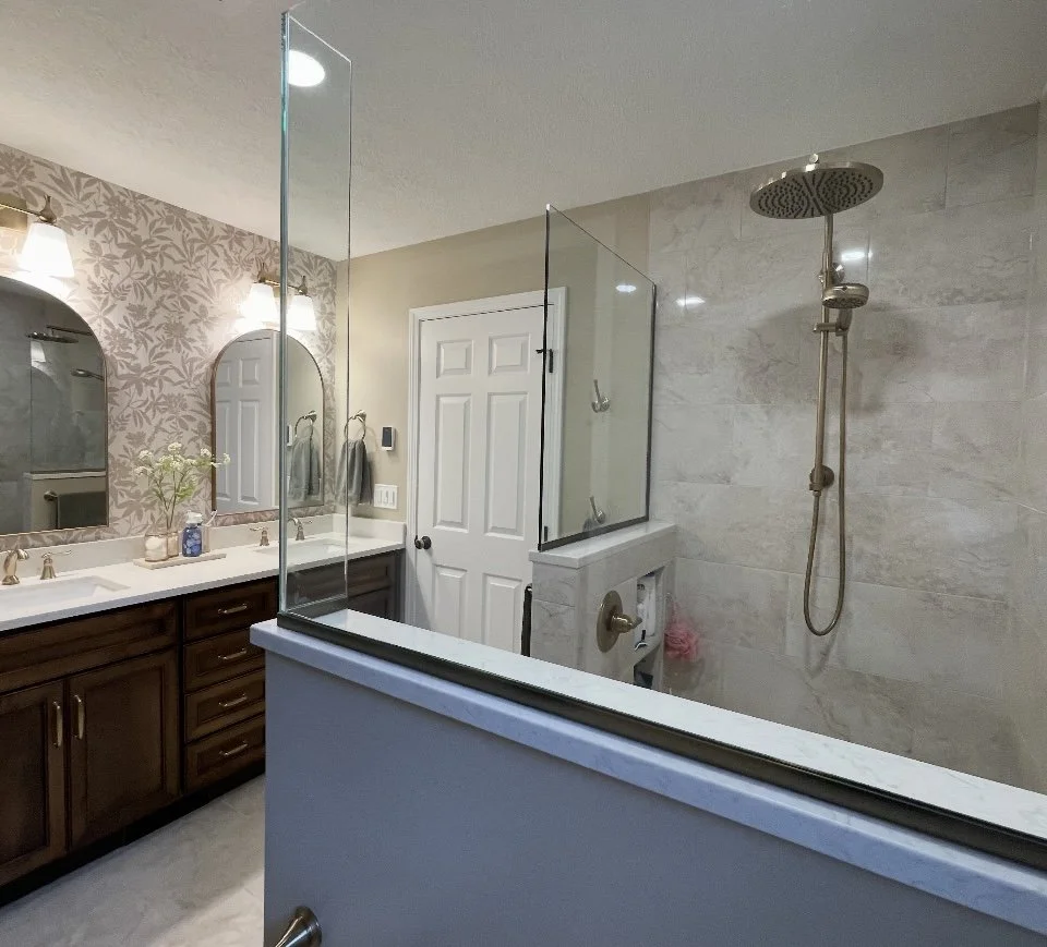 Bathroom with double sink vanity and large mirror, floral wallpaper, and a walk-in shower with glass partition and rainfall showerhead.