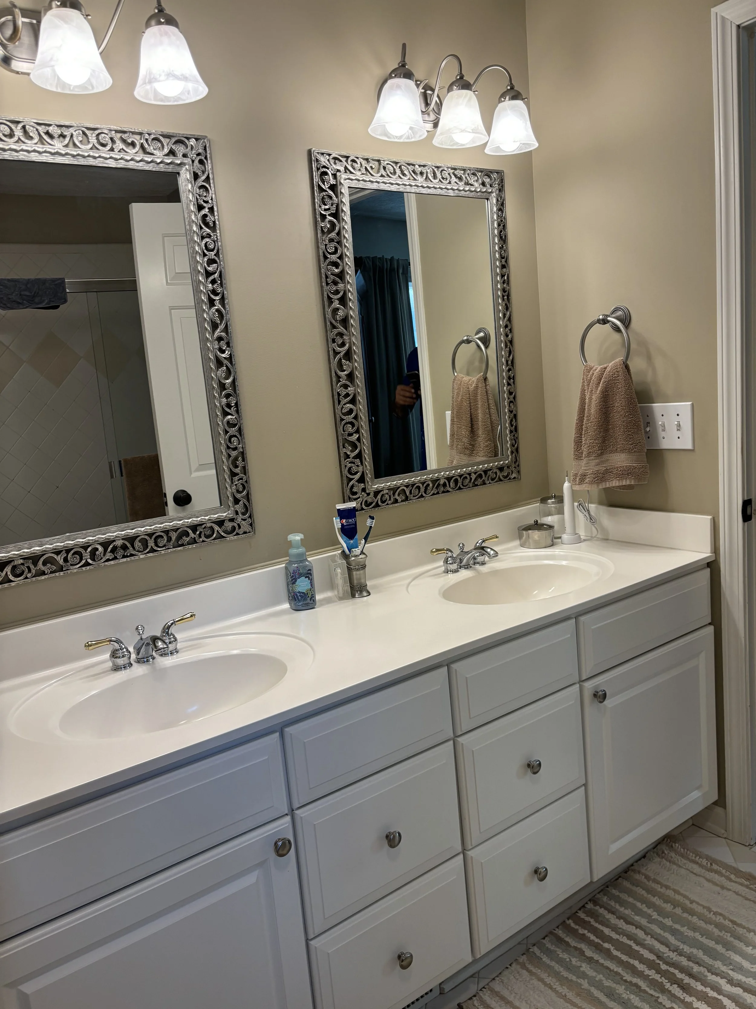 Double bathroom vanity with two sinks, mirrors, and wall-mounted lights in a beige bathroom, with towels on a ring and various toiletries.