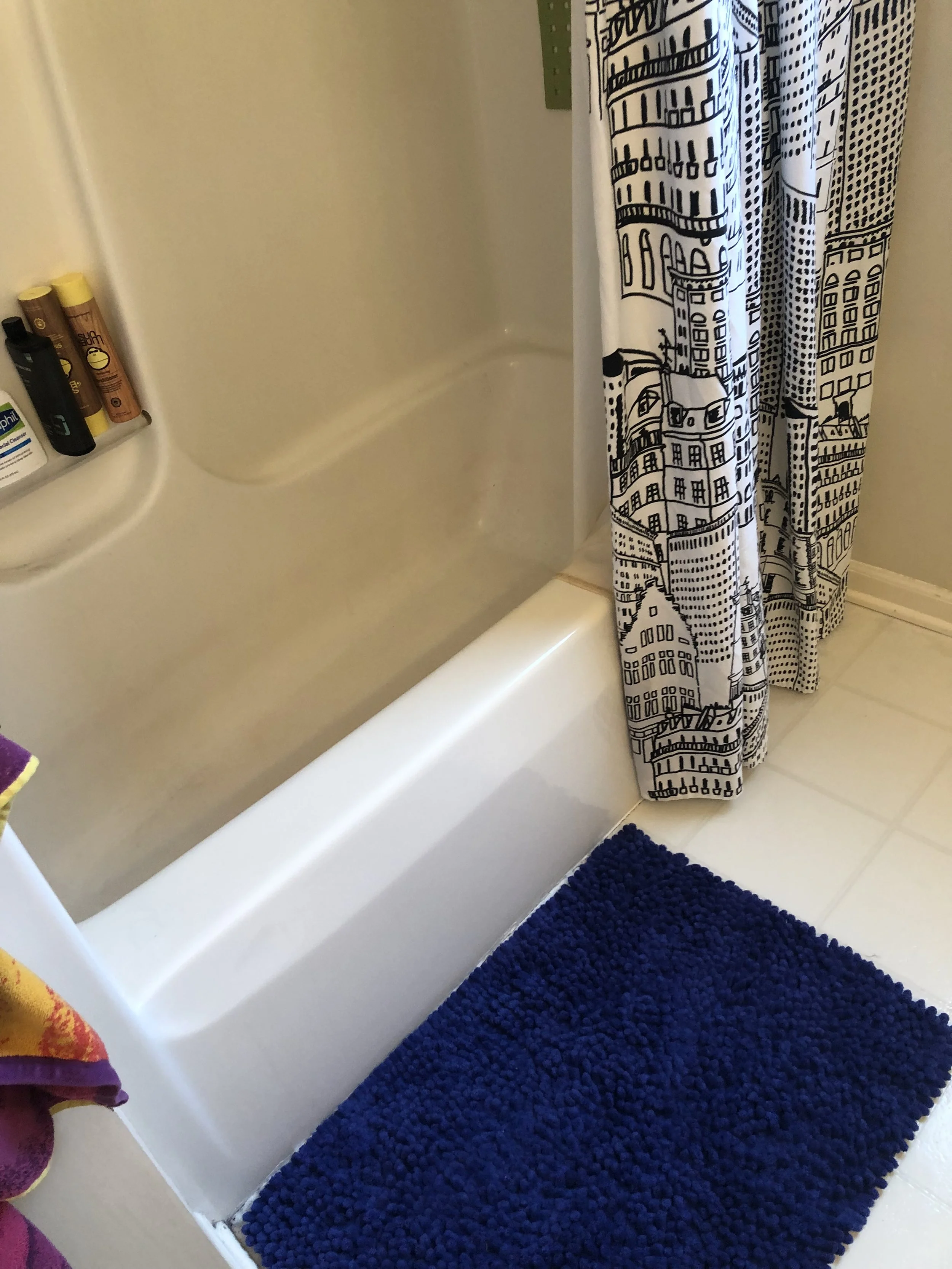 Interior of a bathroom showing a bathtub with a built-in shelf holding various toiletries, a patterned shower curtain, and a blue textured bath mat on the tile floor.