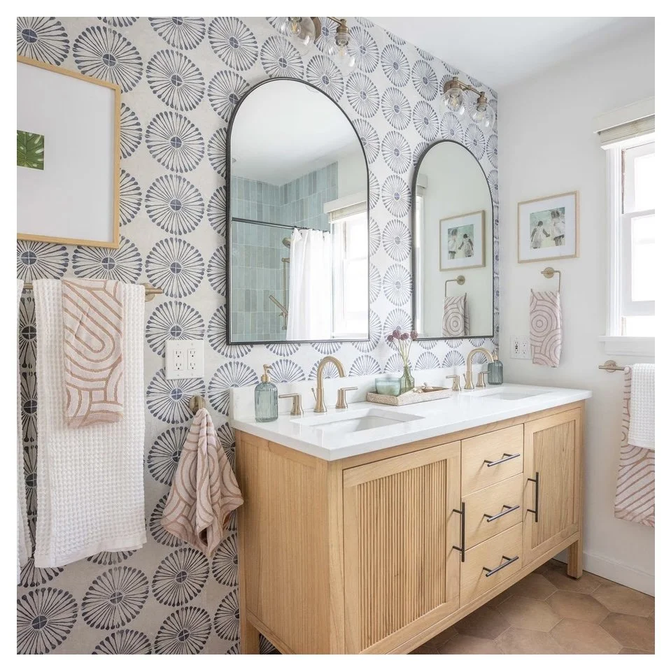 A bathroom with a patterned wallpaper, two oval mirrors, a double sink vanity with wooden cabinets, gold fixtures, and decorated with small vases and framed artwork.