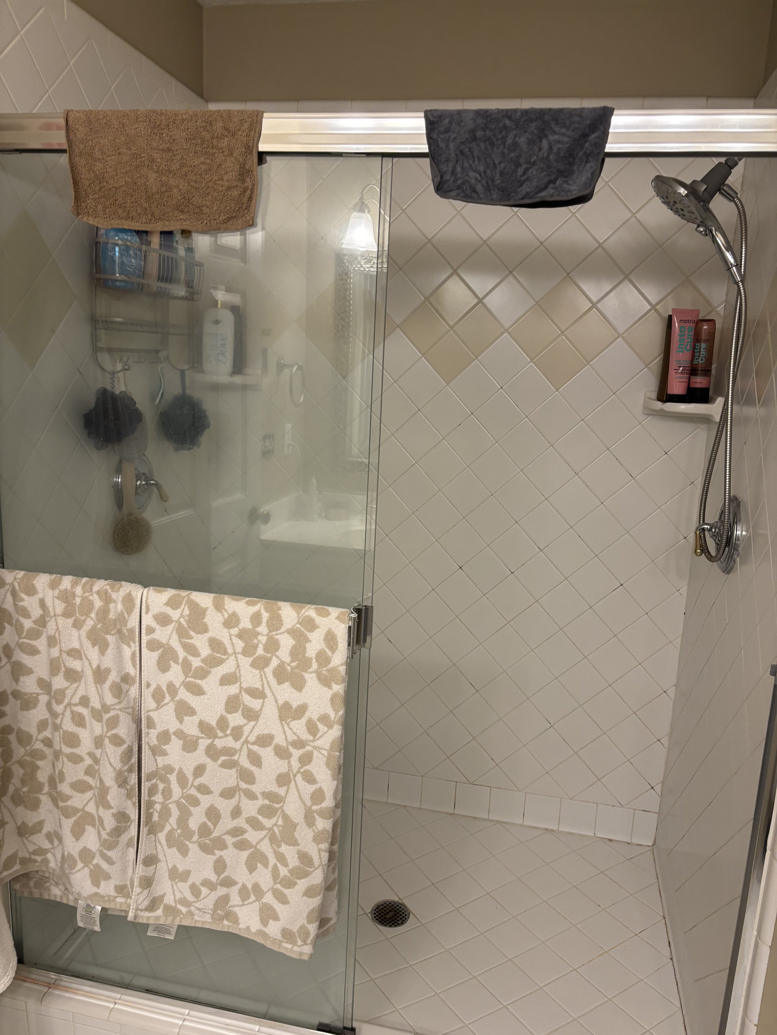 Shower area with beige and white tiles, a glass door with a towel hanging on it, showerhead, and a wall-mounted shelf with shampoo and conditioner bottles.