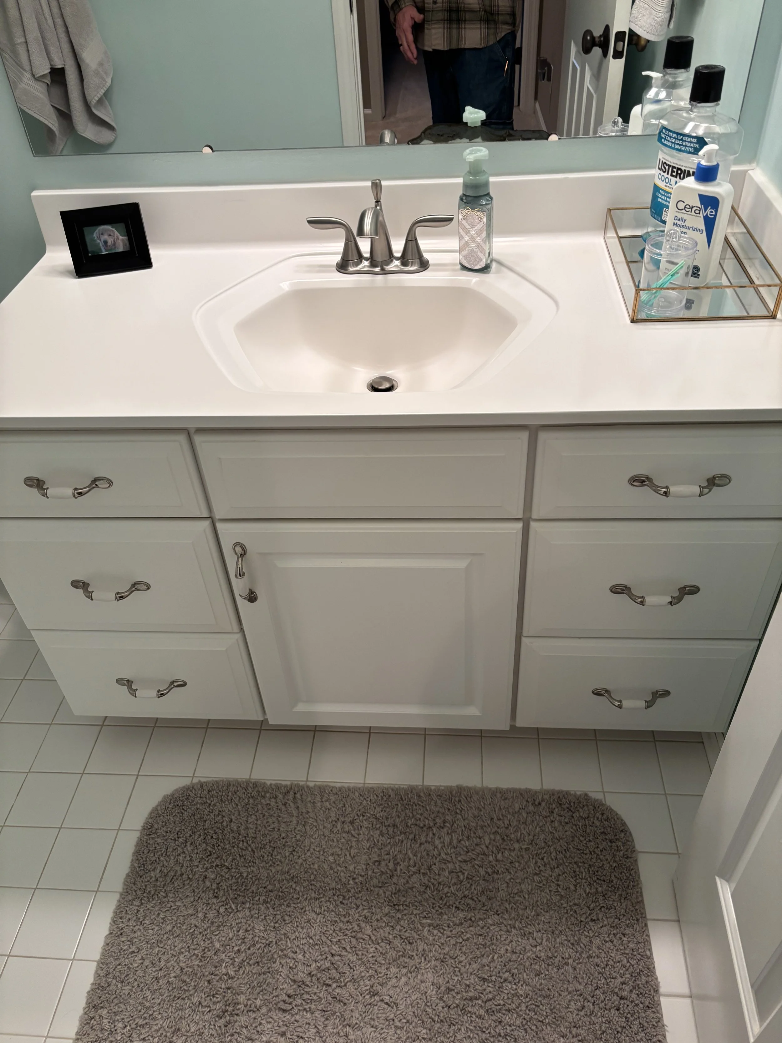 Bathroom vanity with a white countertop, a sink, a silver faucet, a small black framed photo, and a gold tray with hand sanitizer, lotion, and a toothbrush holder. A large mirror reflects part of the person taking the photo. There is a gray bath mat on white tiled floor.