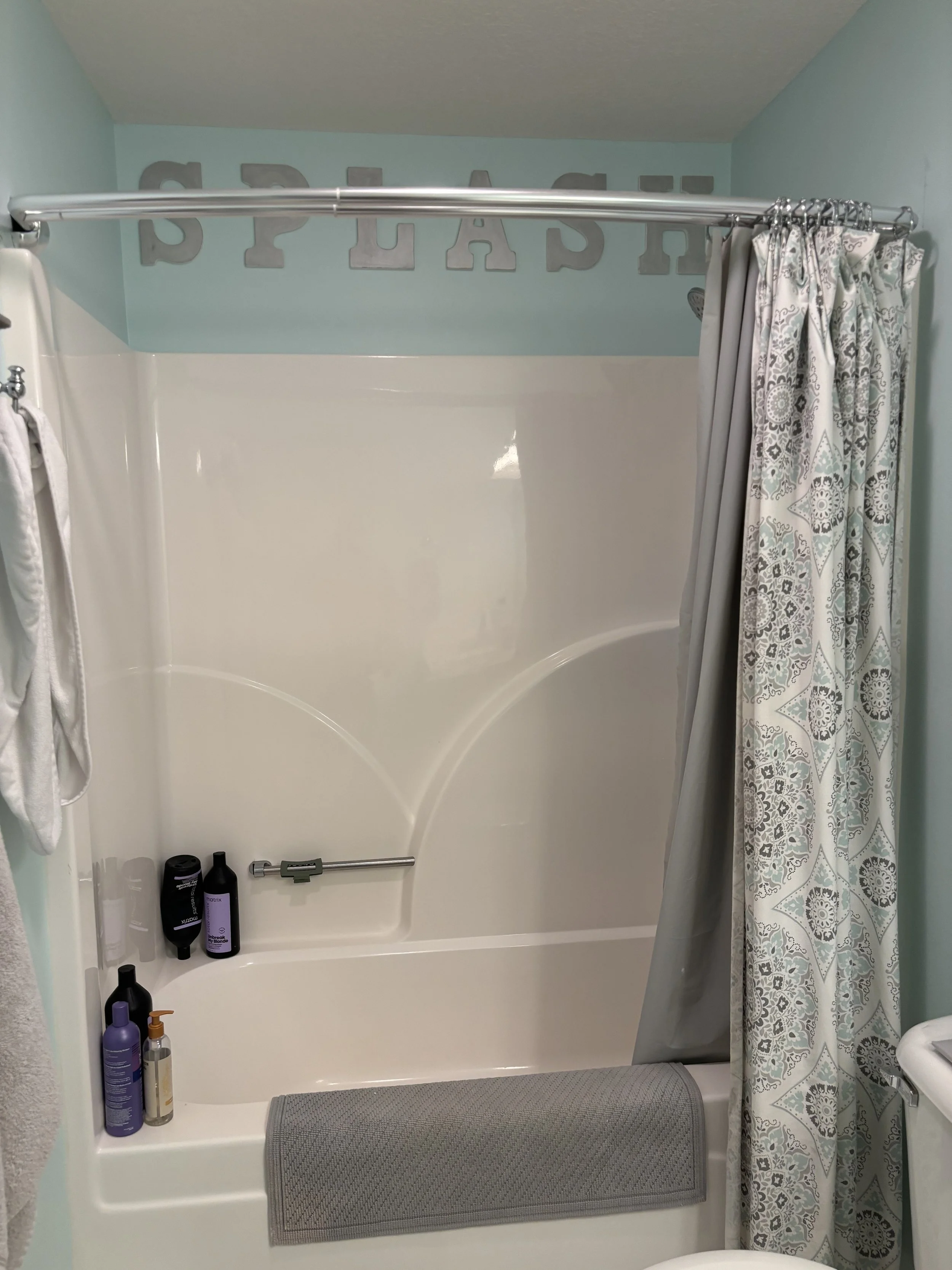 Bathroom with bathtub, shower curtain, light blue wall with decorative letters spelling 'GRA'. Bottles of shampoo, conditioner, and body wash on the tub ledge, towel hanging on hook, gray bath rug at the bath entrance.