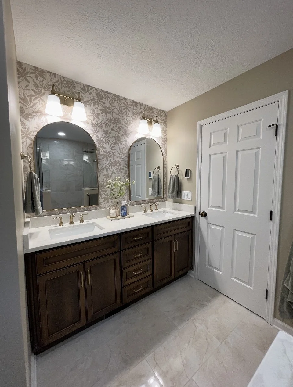 Bathroom with double vanity, dark wooden cabinets, white countertop, two arched mirrors, two wall-mounted light fixtures, beige walls, patterned wallpaper behind mirrors, towels on towel rings, tiled floor, door on the right.
