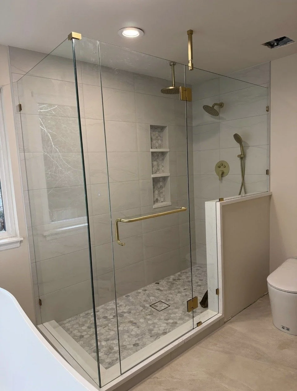 Modern glass shower enclosure with brass hardware, marble niche, and rainfall showerhead, adjacent to a toilet in a bathroom.