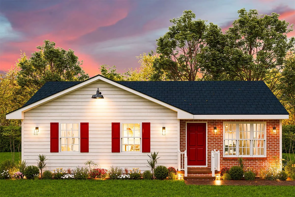 A small house with white siding, red shutters and door, brick accents on one side, surrounded by a garden with plants and flowers, at sunset with a colorful sky and trees in the background.