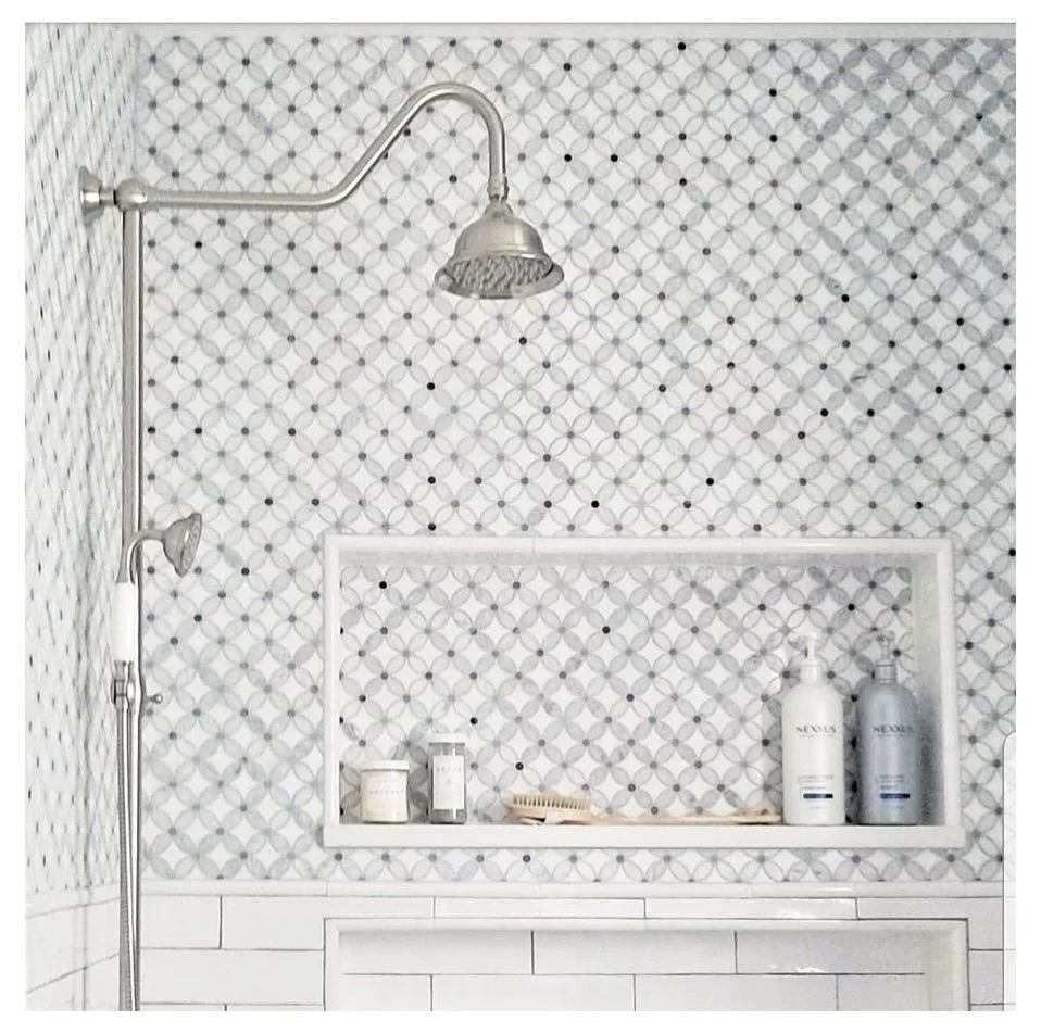 A modern shower area with a patterned tile wall, a large silver showerhead, a smaller handheld shower, and a built-in shelf holding bottles and toiletries.