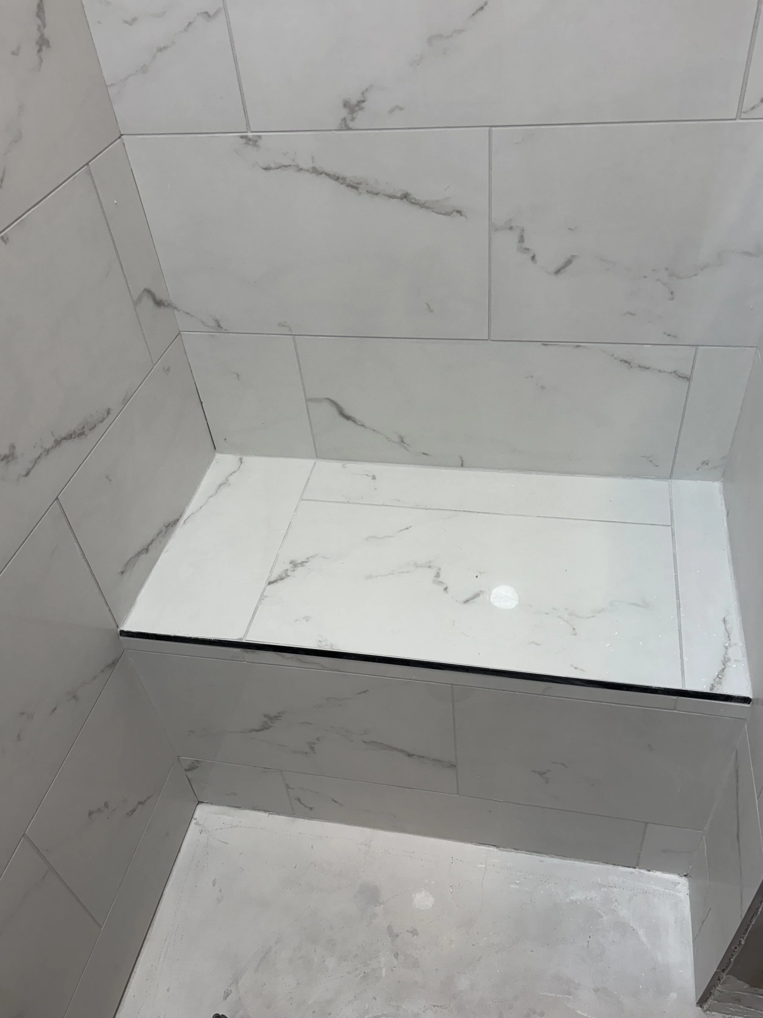 Shower wall and floor with white marble tiles featuring gray veining, including a built-in bench with matching tiles and a drain on the floor.