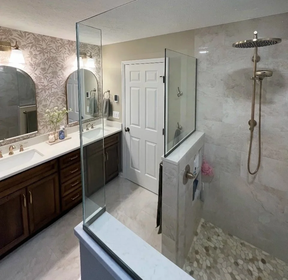 Bathroom with dual sinks and large mirrors on the left, a glass partitioned shower with a rain showerhead on the right, a white door in the center, and decorative wallpaper and lighting.