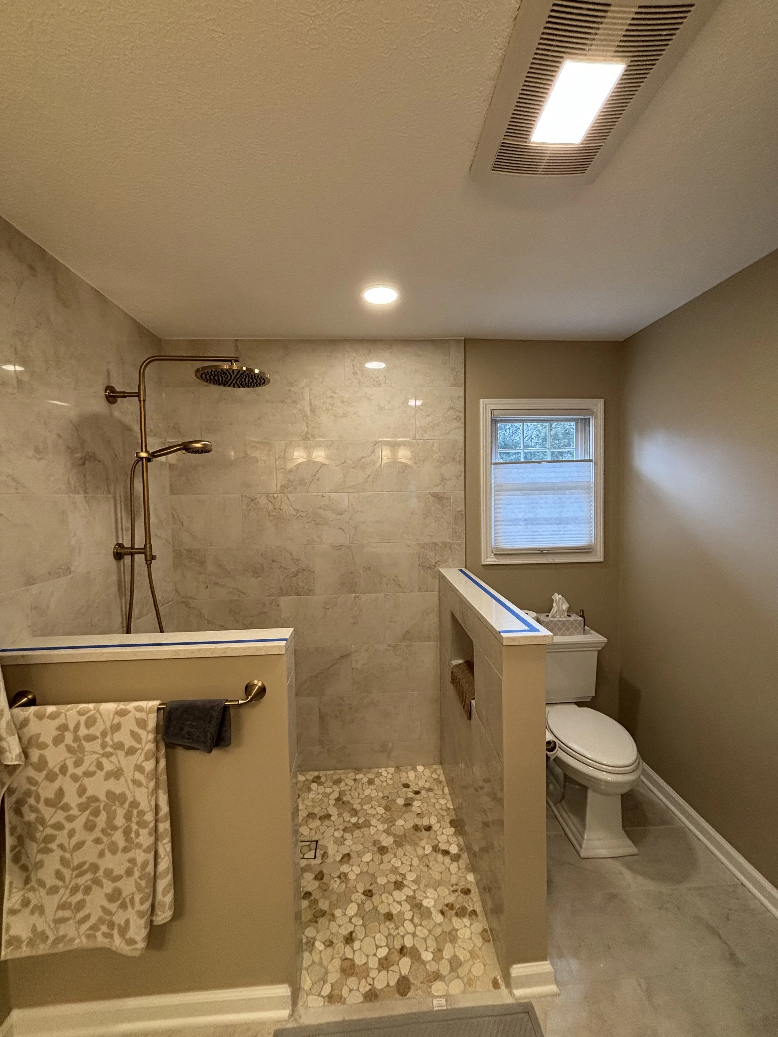 Bathroom with a walk-in shower with beige tile walls and pebble floor, toilet, window, and beige walls.
