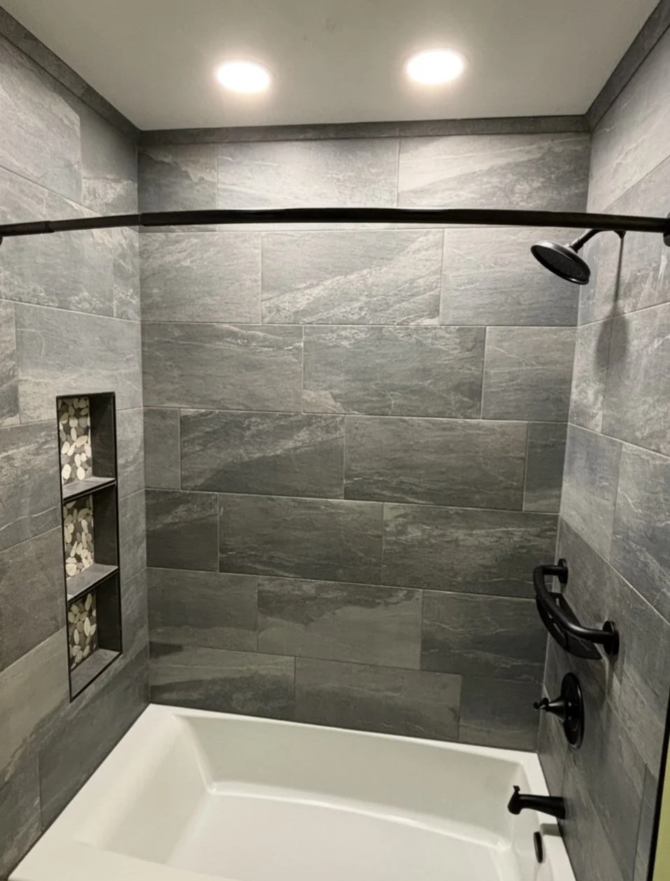 A modern shower with gray tile walls, a black showerhead, and built-in niche with pebble mosaic tiles. The shower has a curved shower curtain rod and black fixtures.