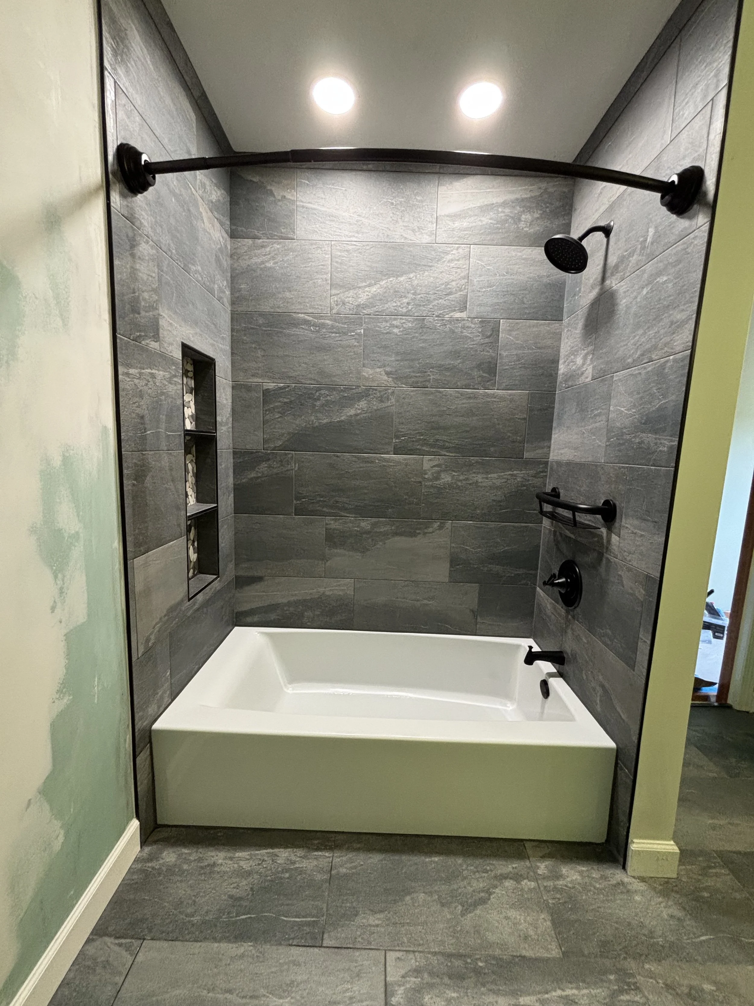 Black showerhead, curtain rod, and fixtures in a modern shower with gray tiles and built-in shelves.