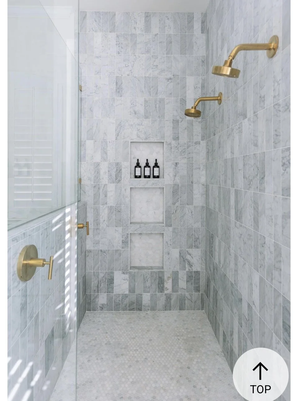 Empty walk-in shower with marble tile walls, three brass shower heads on the right wall, built-in marble niches with three bottles, glass door on the left, and small mosaic tile floor.