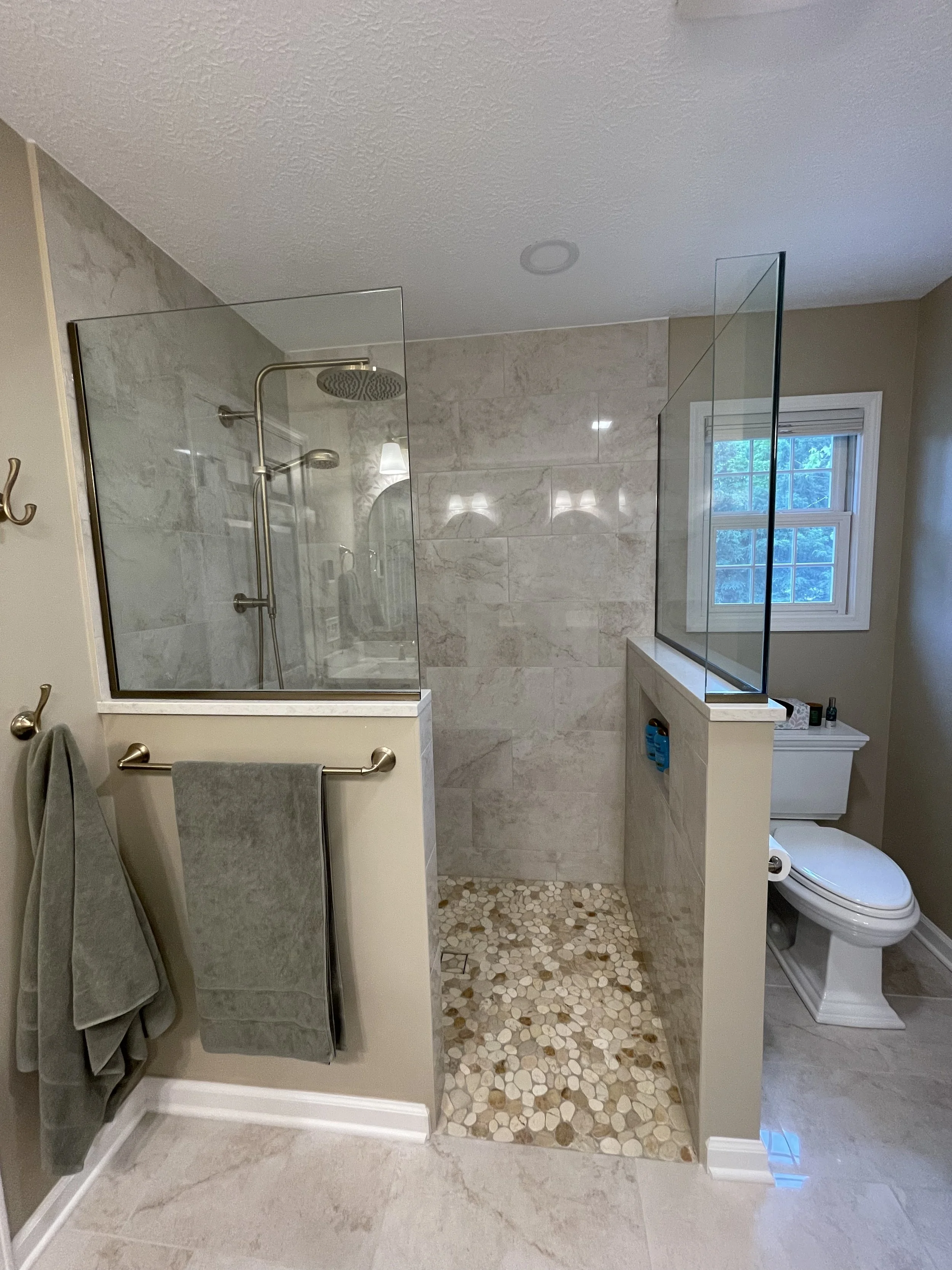 Bathroom with separate shower area enclosed with glass, beige tiled walls, pebble stone shower floor, toilet, window, and towels hanging on a rack.