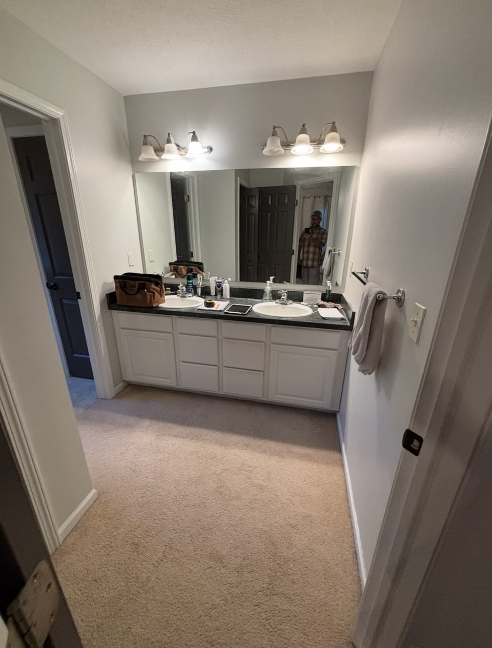 A bathroom with a double sink vanity, mirror, light fixtures, and a person taking a photo in the mirror. Countertop has toiletries and a bag. Towel on a wall hook, beige carpeted floor, doorway on the left, dark doors reflected in the mirror.