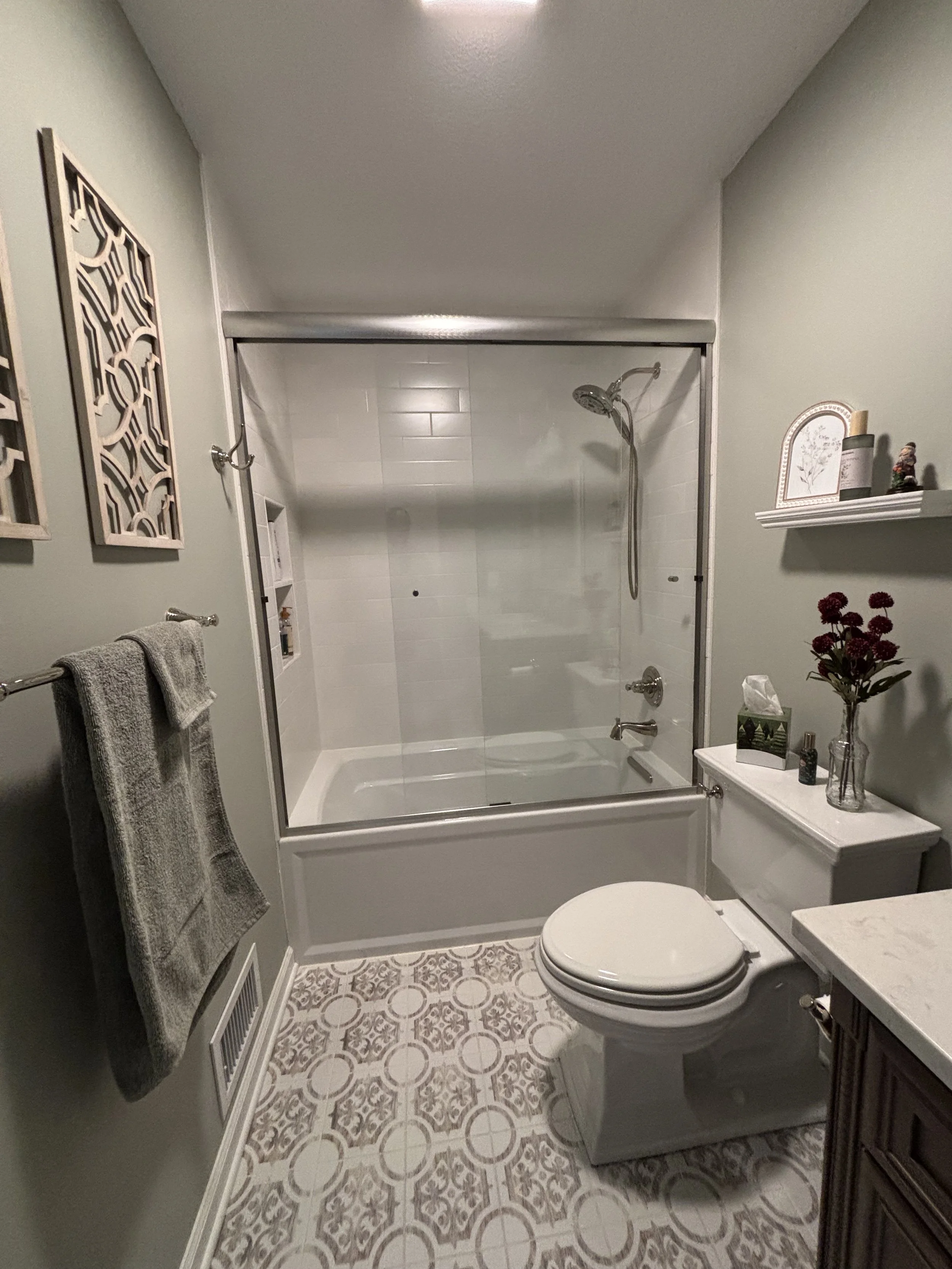 A small bathroom with a bathtub and shower enclosed by glass doors, a toilet, and a vanity with a countertop. The walls are painted light green, and decorative wall art and a shelf with small items are present. The floor has patterned tiles.