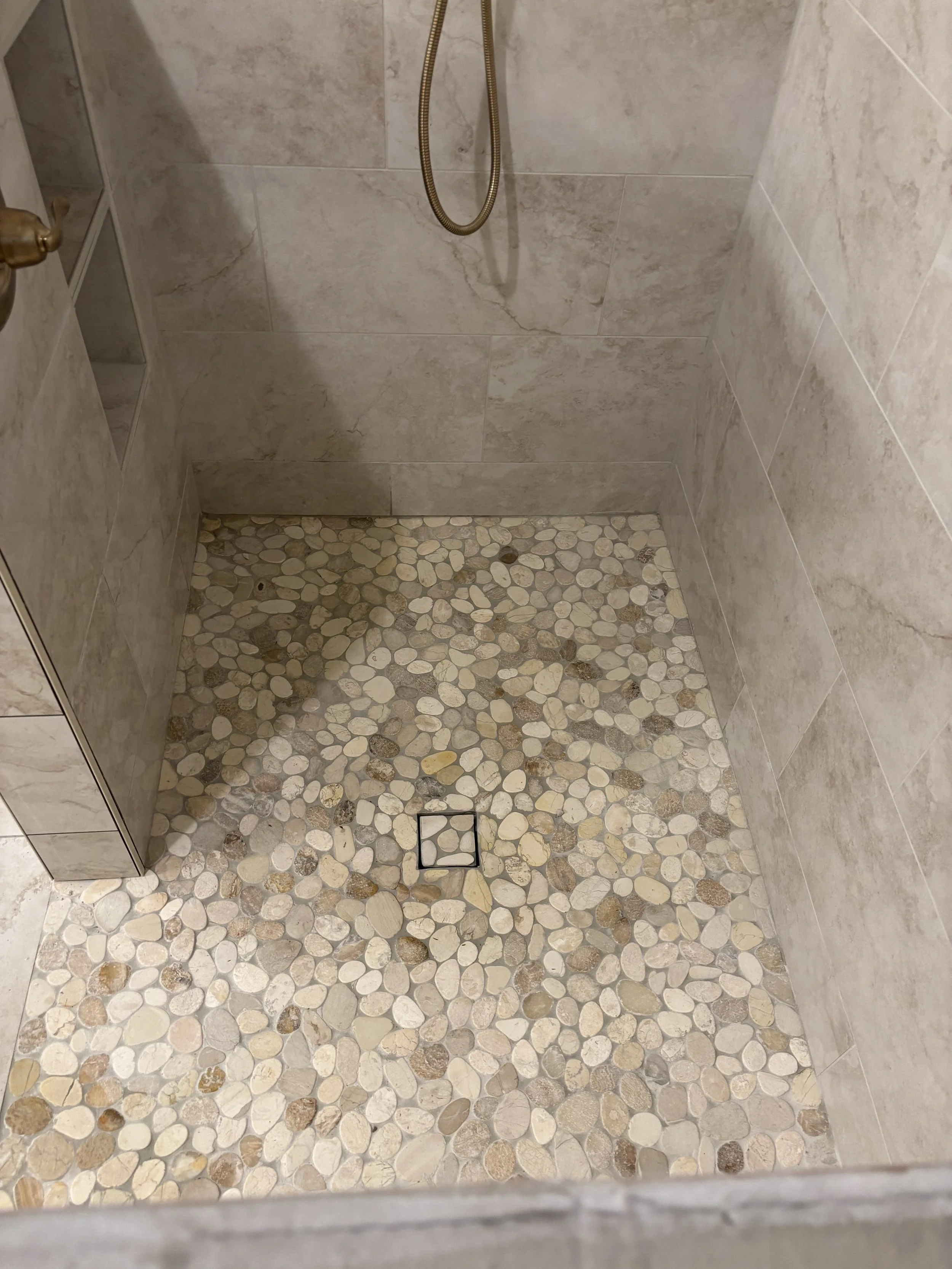 Inside a shower with beige tiled walls and a pebble stone floor with a square drain in the center.