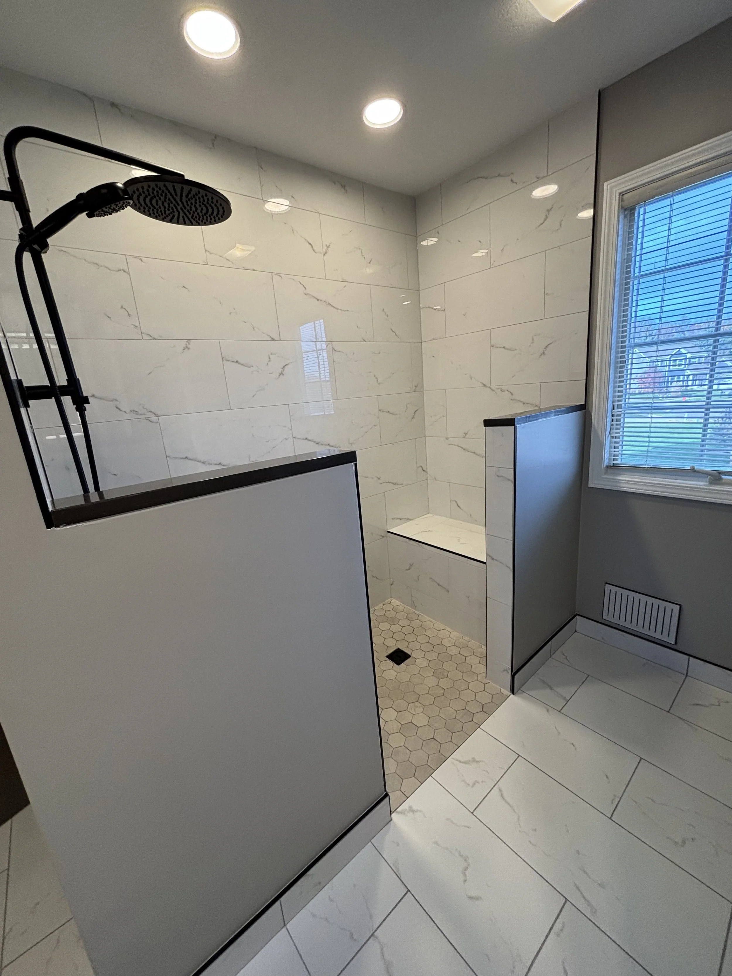 Modern bathroom shower area with marble tiles, a built-in bench, a rainfall showerhead, and a window with blinds.
