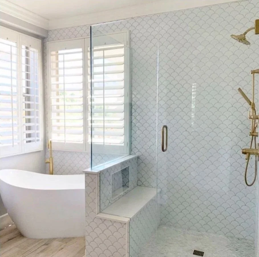 Modern bathroom with white freestanding bathtub, large window with shutters, glass shower enclosure with gold fixtures, patterned white tile walls, and light-colored wood floors.