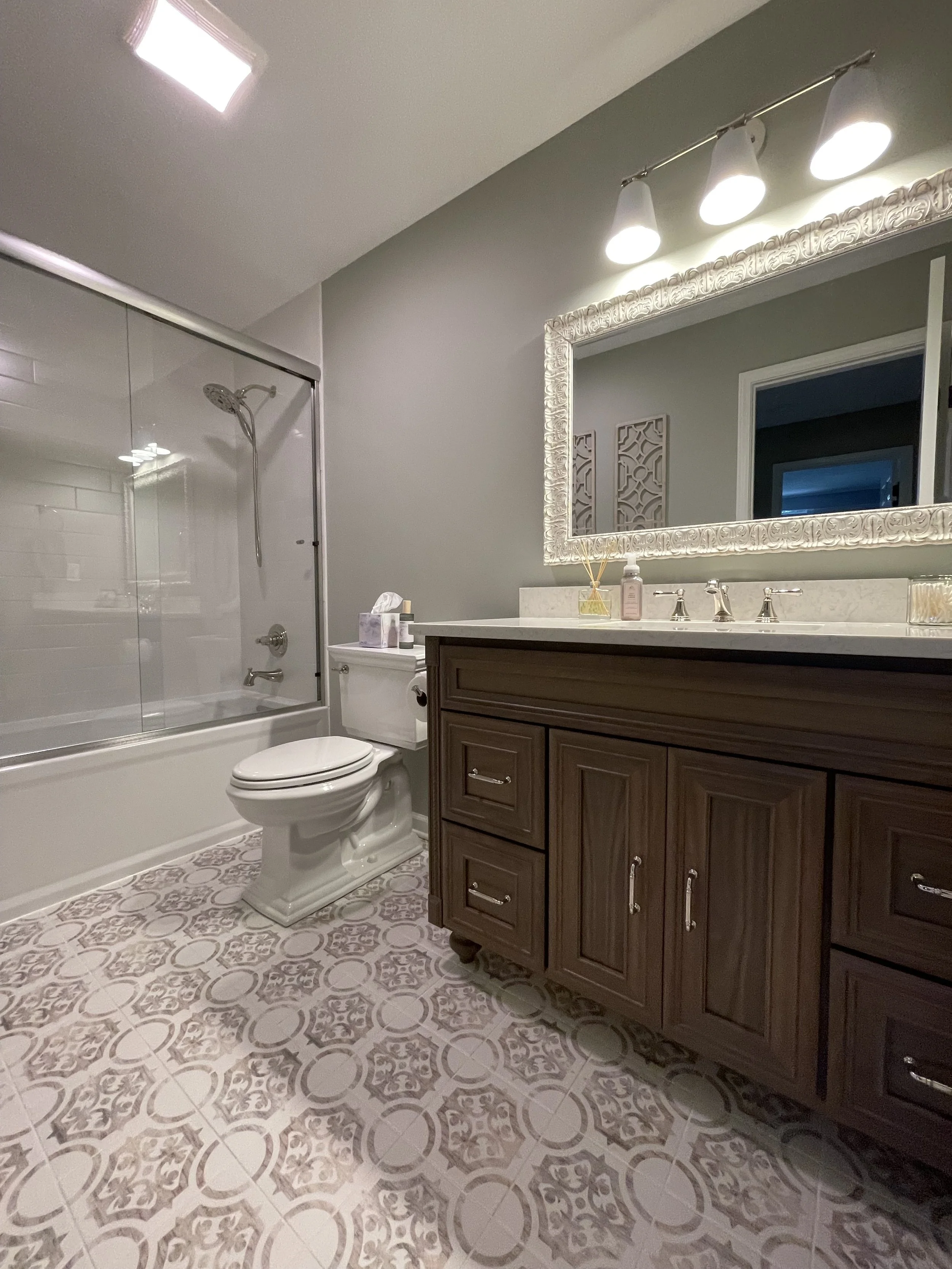 A bathroom with a bathtub and shower, a toilet, a wooden vanity with a white countertop, a large decorative mirror, and modern lighting fixtures.