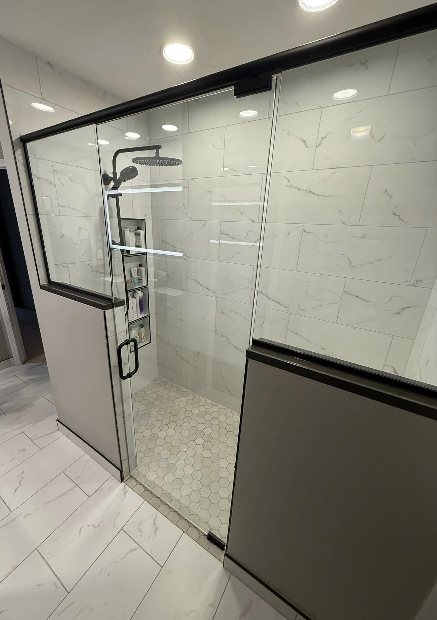 A glass shower enclosure with a black handle, marble walls, a rainfall showerhead, and a built-in shelf with toiletries inside.