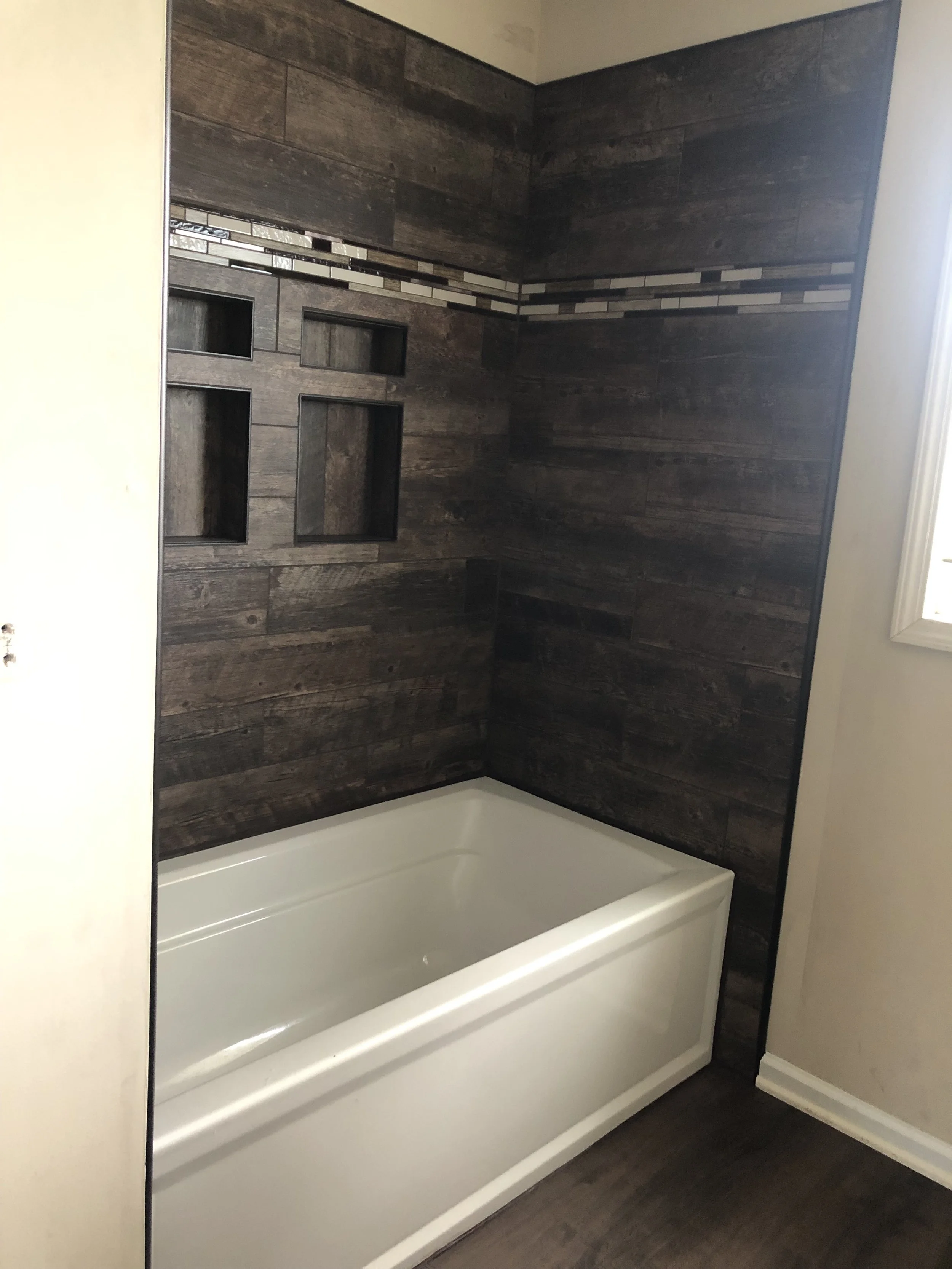 Bathroom with a white bathtub, dark wood tile walls, a decorative tile strip, built-in wall niches, and a window.