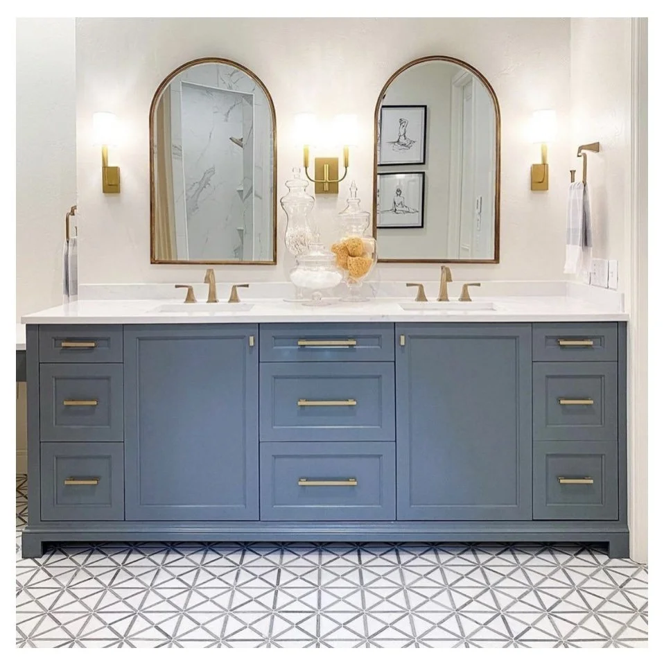 Bathroom vanity with two mirrors, three double light fixtures, and a blue cabinet with gold hardware. The countertop has jars and a towel.