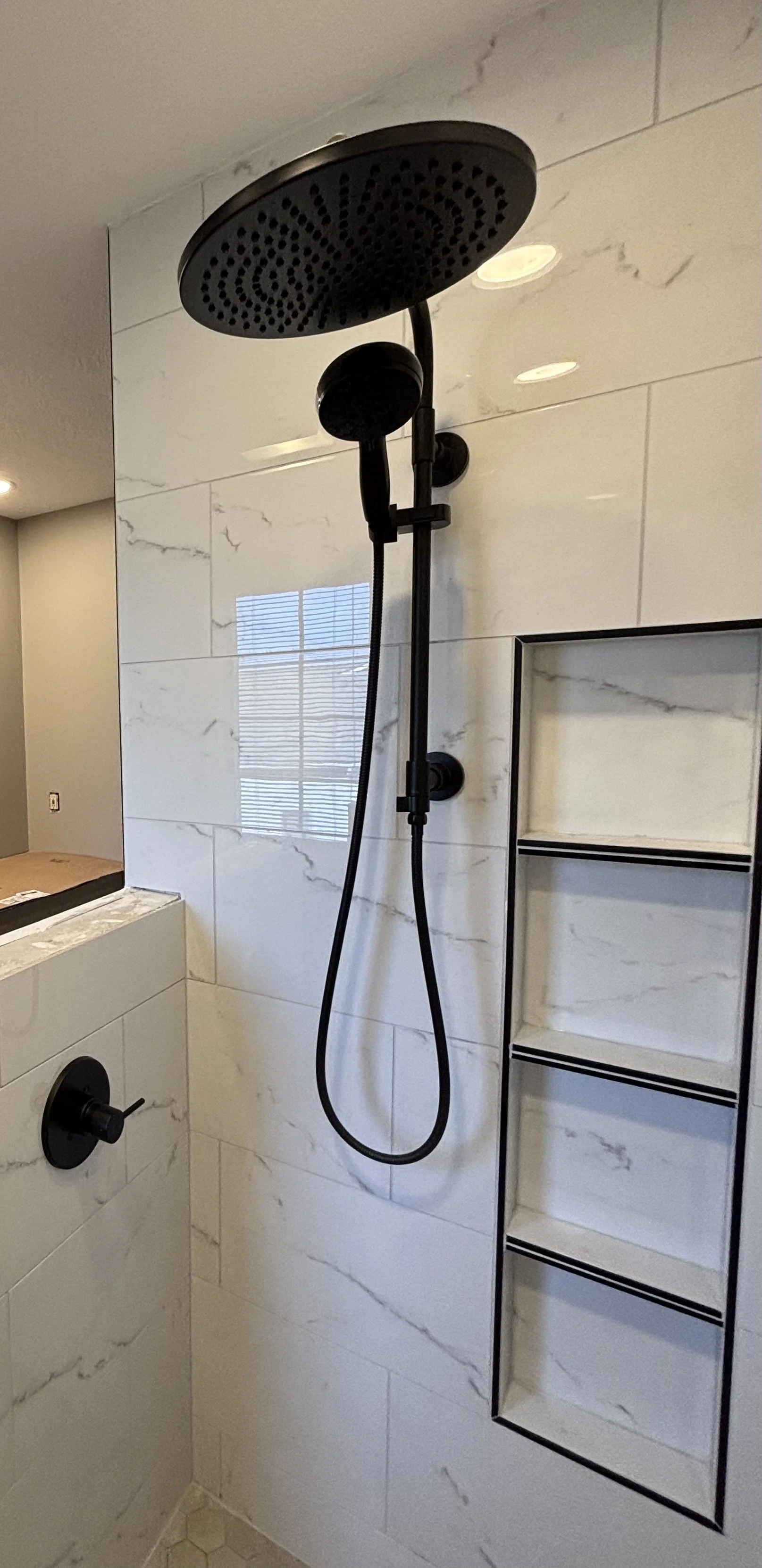 Black showerhead and handheld shower attachment mounted on a white marble wall in a bathroom shower area with built-in shelves.