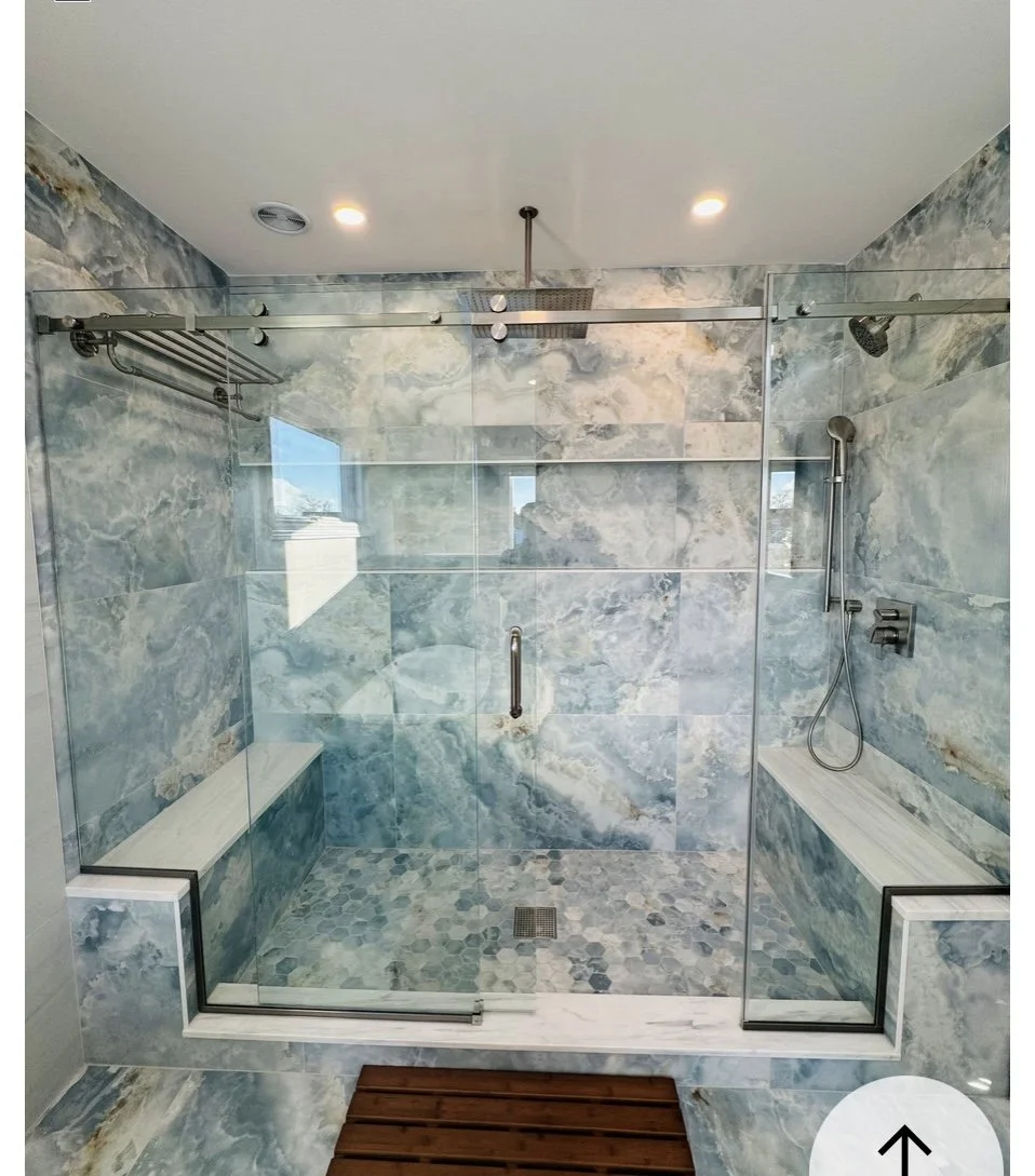 Modern walk-in shower with glass enclosure, marble tiles, built-in benches, rain showerhead, and handheld shower, with a wooden bath mat outside.