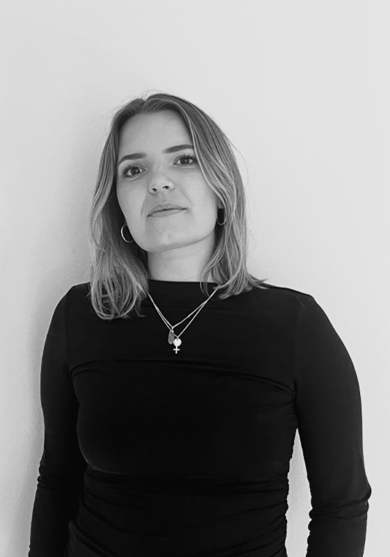 Kvinna med axellångt hår, iförd svart långärmad topp och halsbandsmycken, står framför en vit vägg.