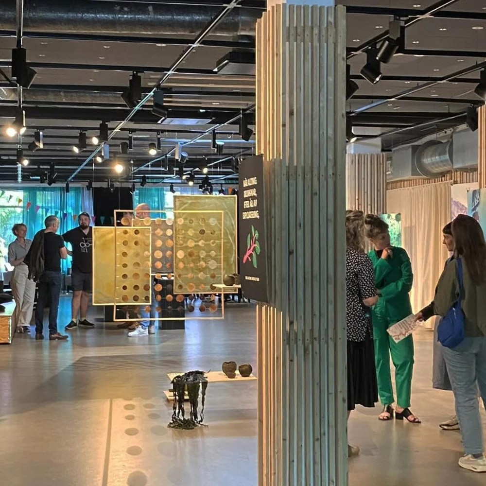 Besökare i en utställningslokal under Souther Sweden Design Days 2024. Fyra personer står och pratar till höger, några andra i bakgrunden diskuterar. Utställningen har träramar och konstnärliga element.
