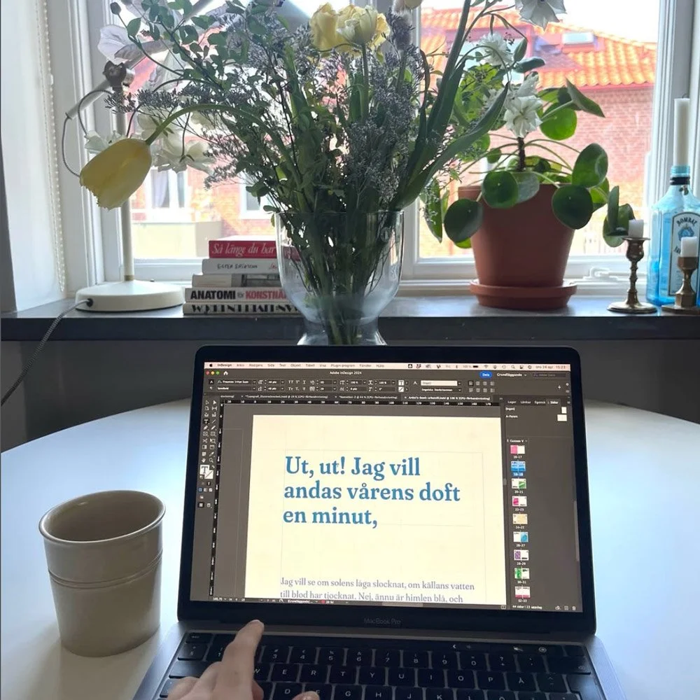 Ett vitt runt bord med en öppen dator, ett kaffe i en vit mugg, en blomvas med blommor, ett fönster med utsikt över tak, en växt i kruka, några böcker och några ljusstakar på fönsterkarmen.