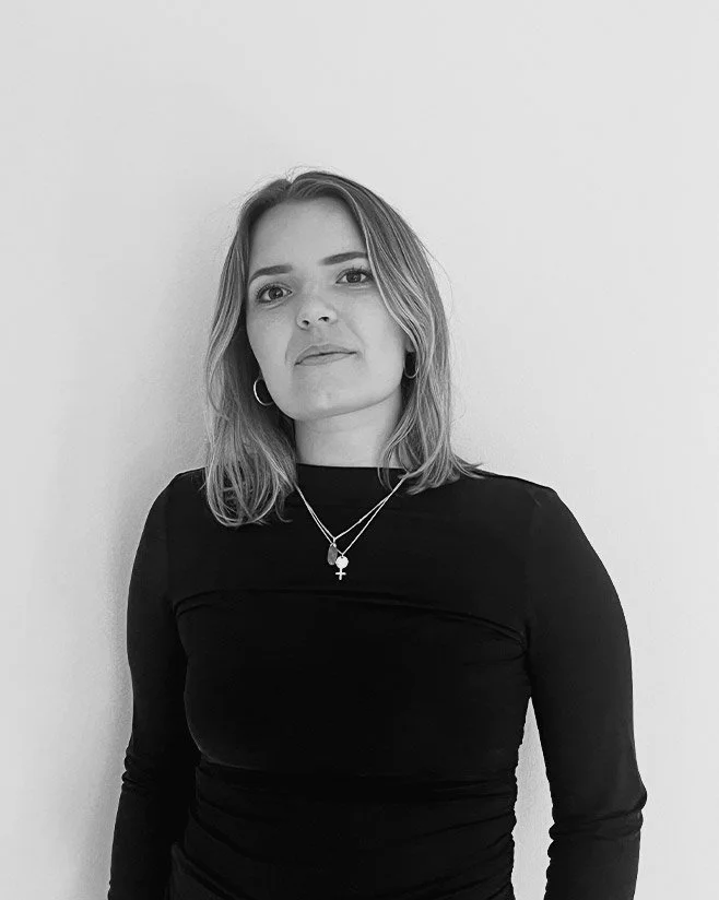 En ung kvinna, som heter Alma Ahnlide med axellångt hår, iförd svart tröja och halsband, står mot en vit bakgrund.