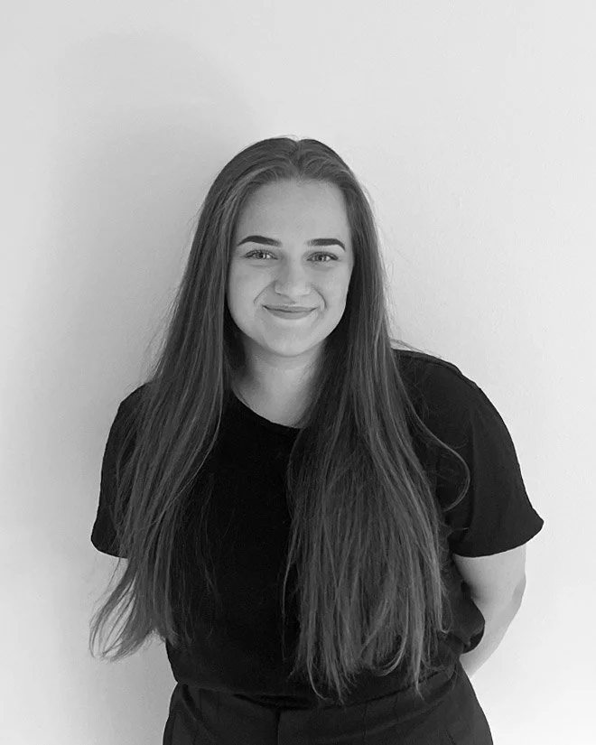 En ung kvinna, som heter Fikrete Meta, med långt, mörk brunt hår som ler mot kameran, bär en svart t-shirt och står framför en ljus vägg.