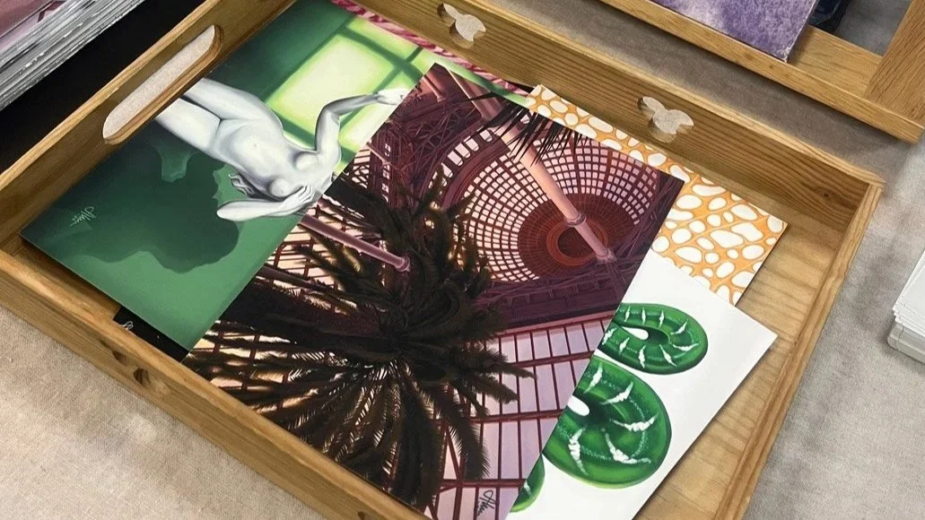 Flera konstverk och fotografier i en träskål, inklusive en illustration av en naken människa på ett grönt bakgrund, n illustration av ett tak med en cirkulär ljusstruktur, en illustration av ett palmtträd, och ett konstverk med gröna blad.